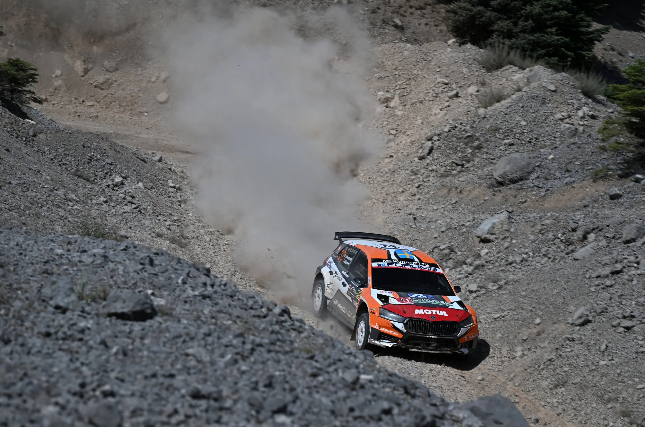 A rally car racing on a dirt road with dust and rocks in a rugged mountainous landscape.