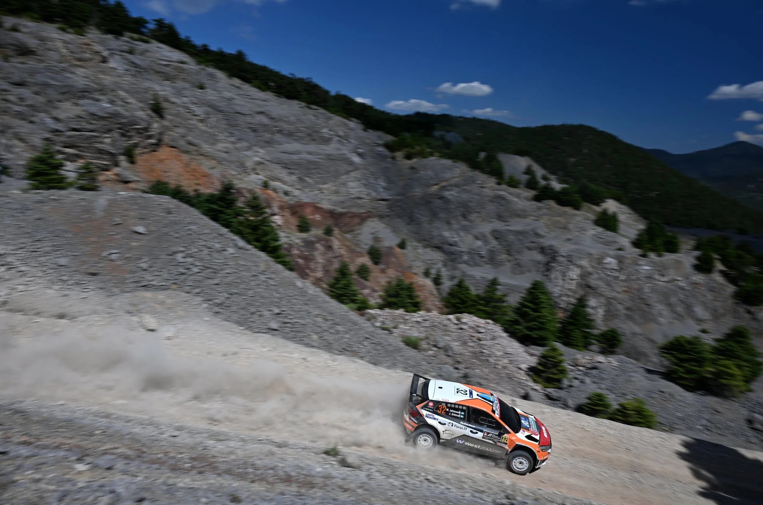 A rally car speeds down a dirt mountain road, kicking up dust behind it, with rugged, rocky mountain terrain and green trees under a clear blue sky in the background.