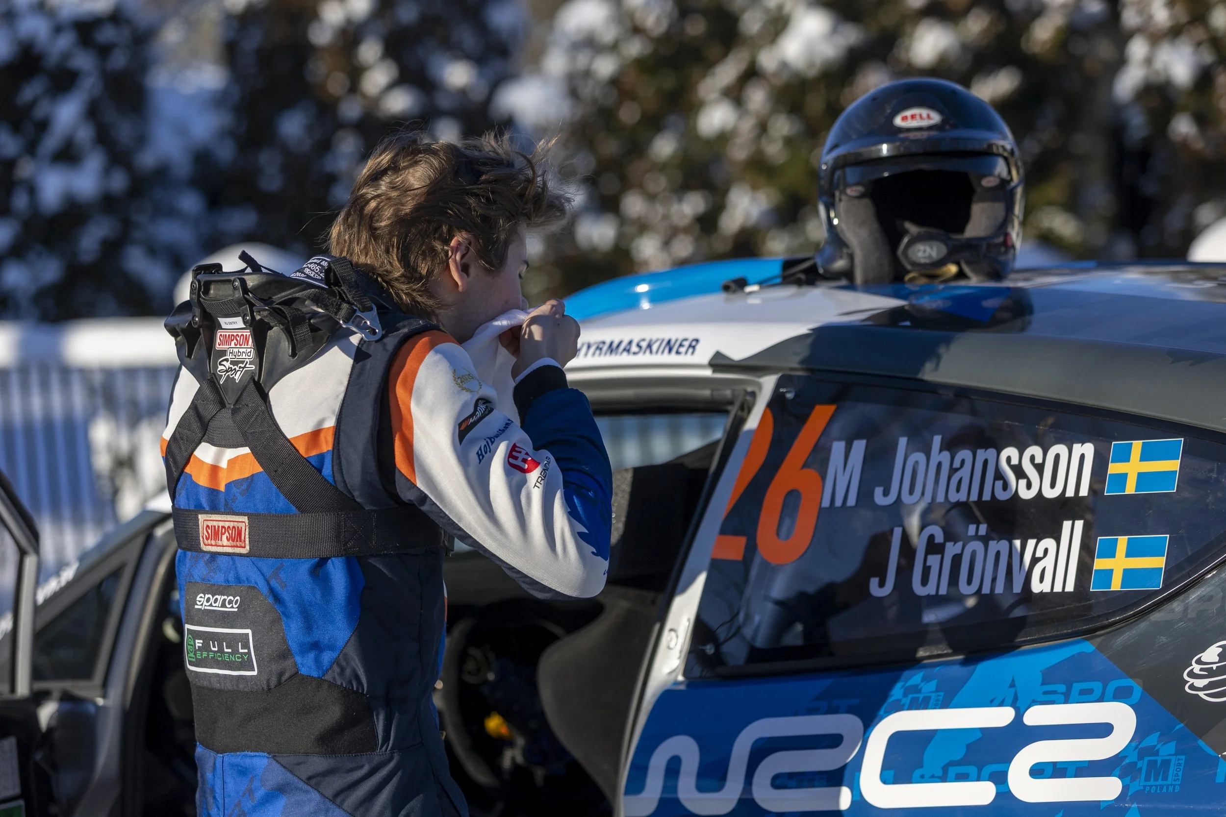 A race car driver with a racing suit is wiping their face with a cloth next to a race car, with a helmet placed on the roof. The car shows the name M. Johansson and Swedish flags.