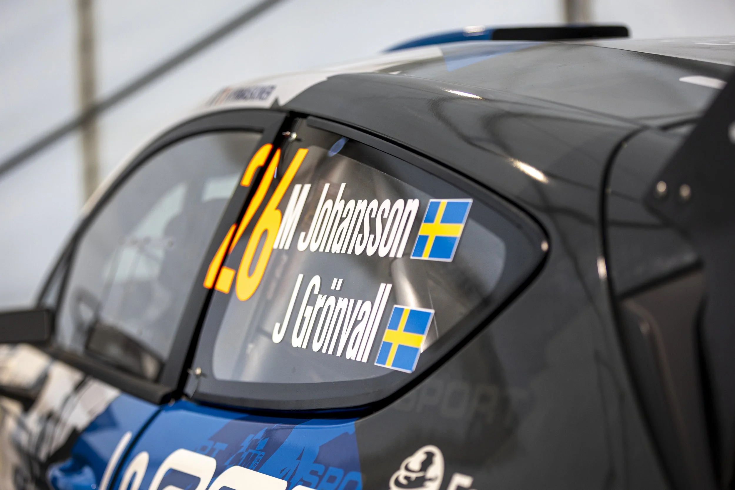 Close-up of a race car's windshield with sponsor decals including a Swedish flag, and the number 16, with a blurred background.
