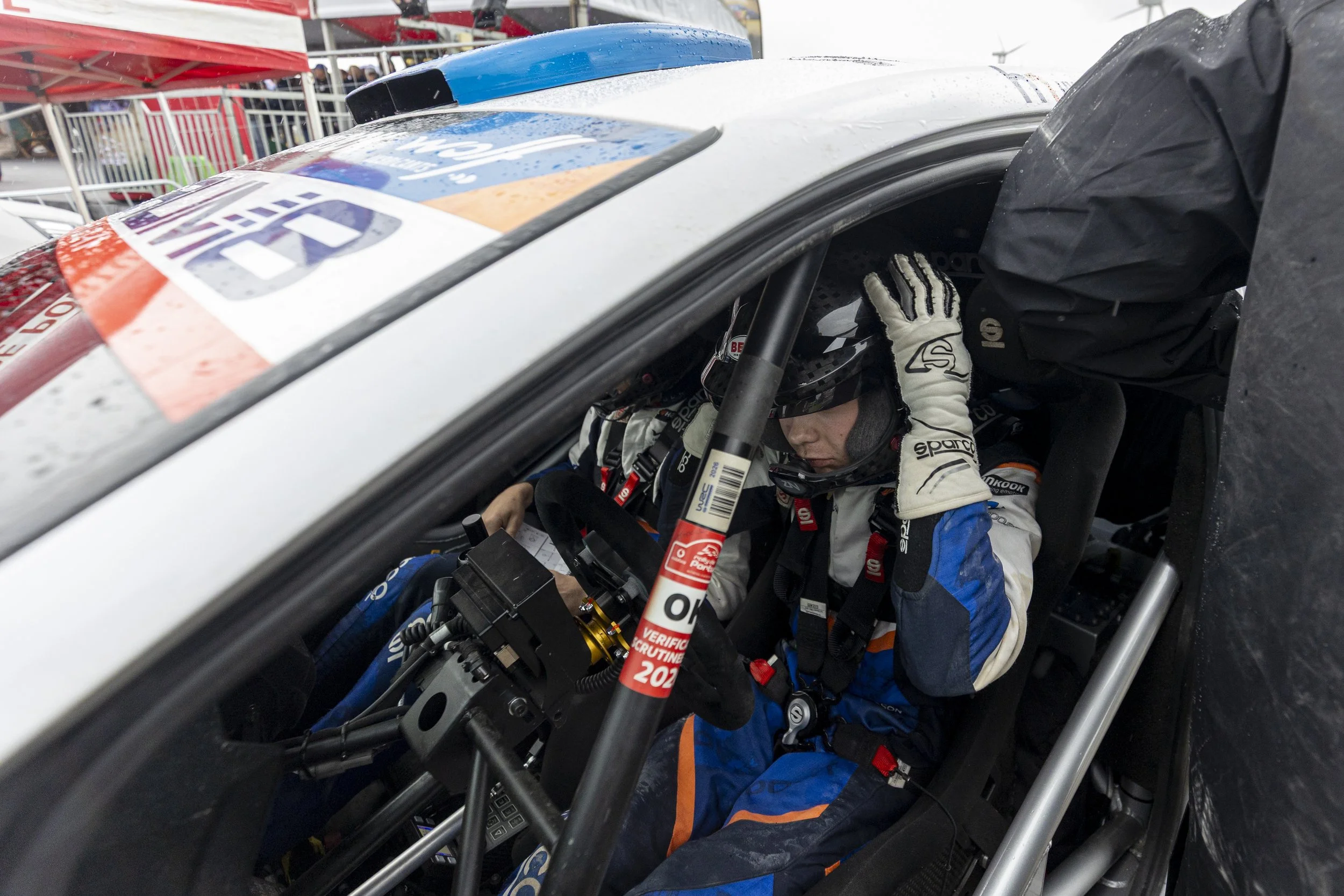 Race car driver in racing suit and helmet sitting inside a race car, looking down, with wet weather conditions and pit stop area visible outside.