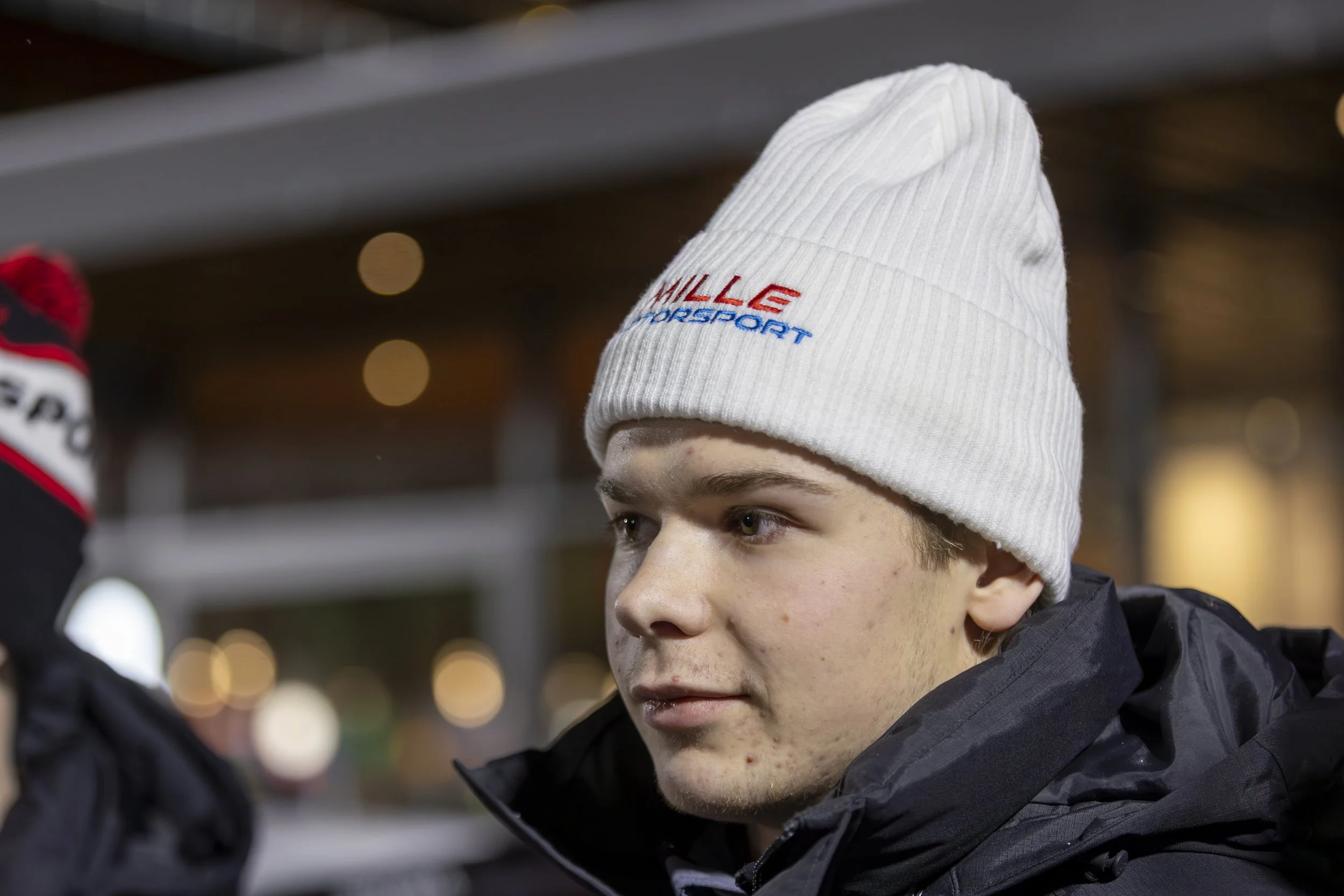 Close-up of a young man wearing a white knit beanie with the words 'MILLE Motorsport' embroidered on it, looking to the side in a nighttime environment.