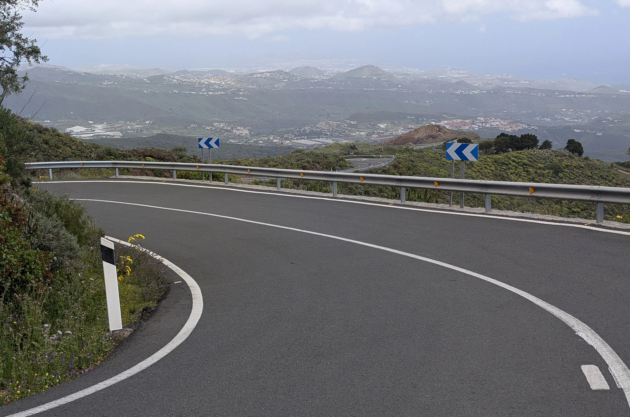 Curving mountain road with guardrail and blue directional signs, overlooking a landscape of rolling hills, scattered buildings, and distant mountains under a cloudy sky.