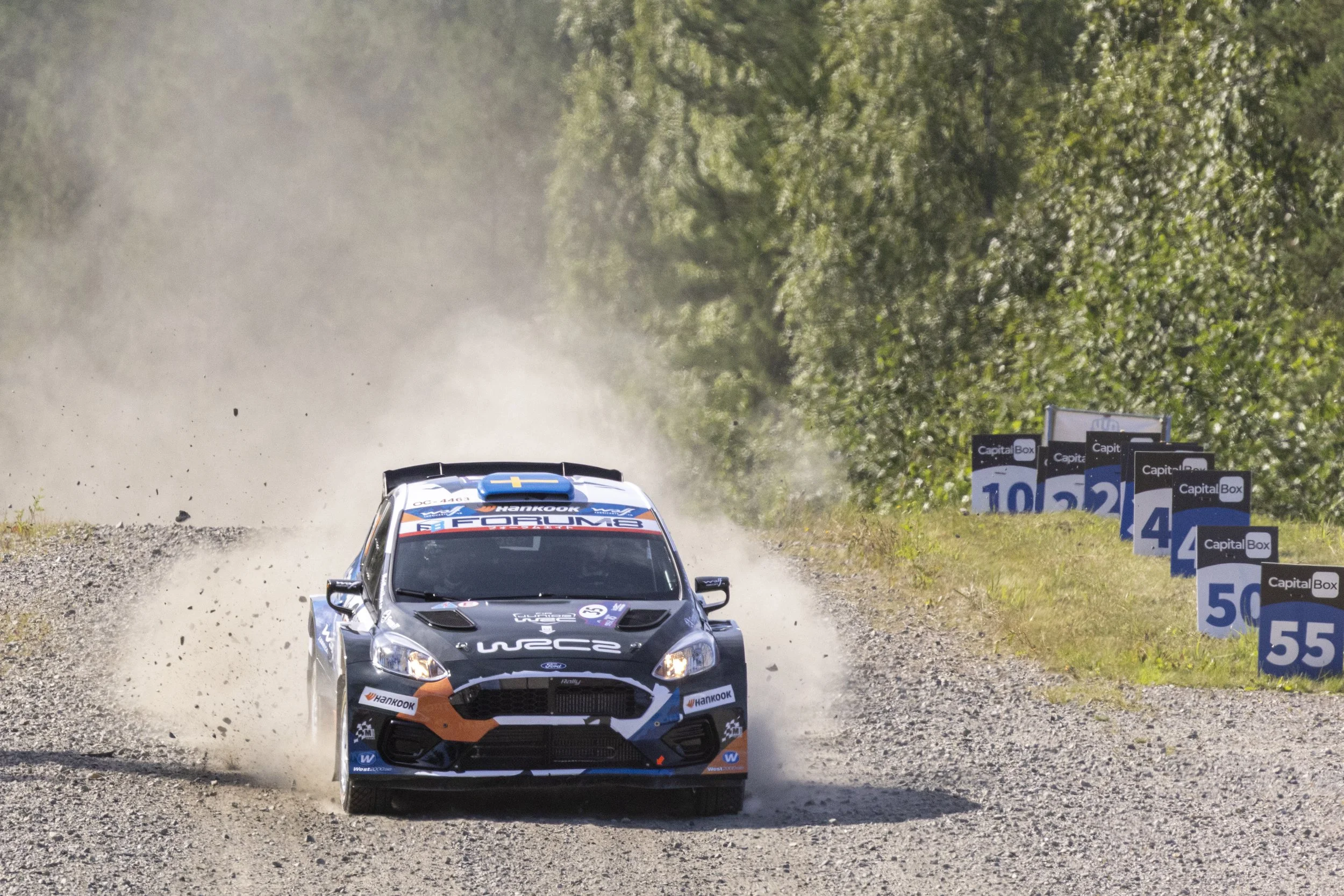 A rally car racing on a gravel road during a motorsport event, kicking up dust with trees lining the sides of the course and speed limit signs visible.