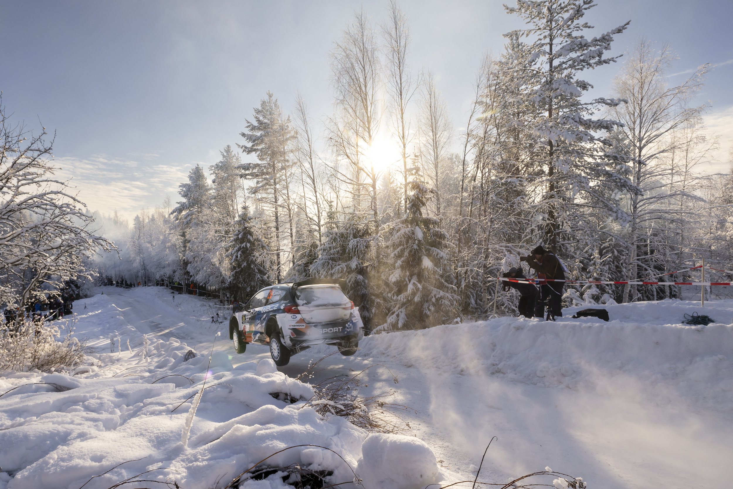 A snowy forest scene with the sun shining through tall trees. A rally car is airborne, jumping over snow-covered terrain. Several people are gathered in the background near the trees, and there are rally officials or photographers near the jump, capt