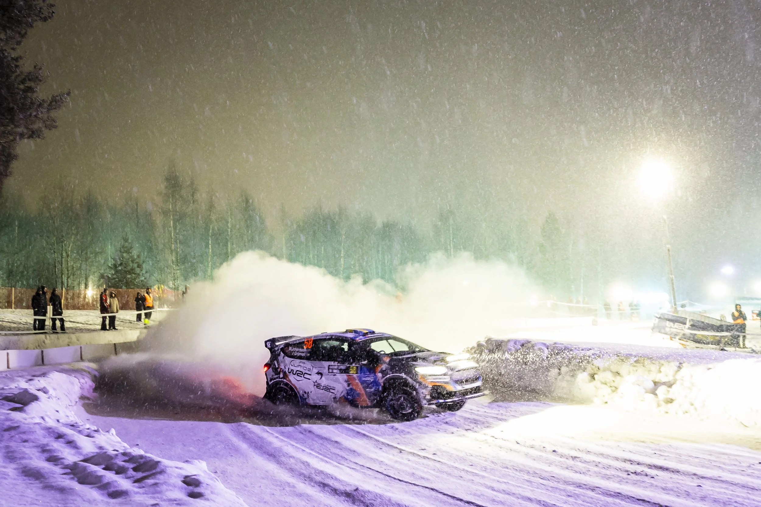Nighttime scene of a snowy rally car race with a car in mid-slide, snow flying, and spectators watching behind a safety barrier. Bright lights illuminate the snowy track and trees in the background.