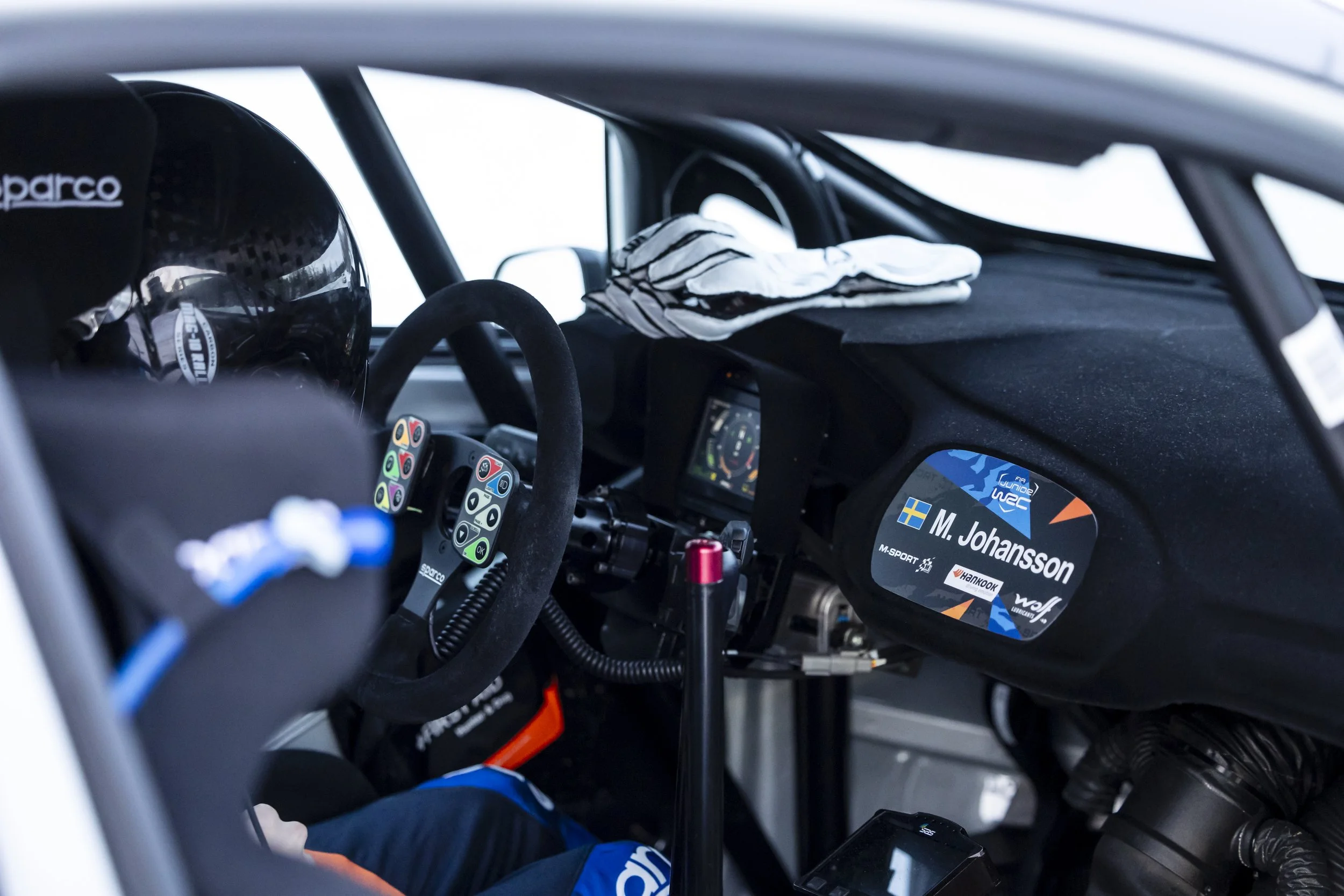 View of the interior of a race car with a helmet on the driver's seat, racing gloves on the dashboard, and a digital display showing the name 'M. Johansson' and Swedish flags.
