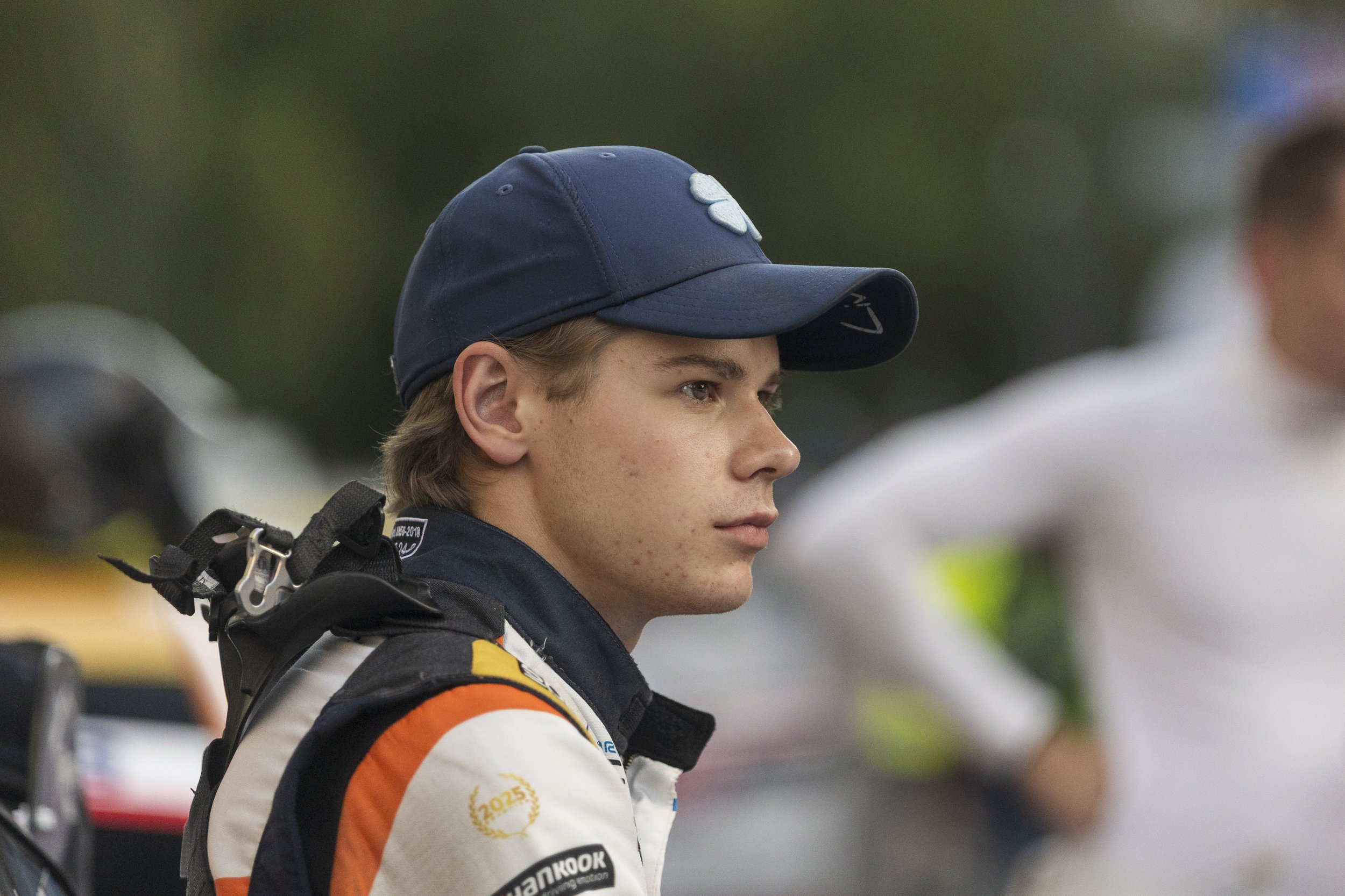 A young male race car driver in racing gear, including a white suit with orange and black accents, wearing a blue cap, standing outdoors at a racing event.