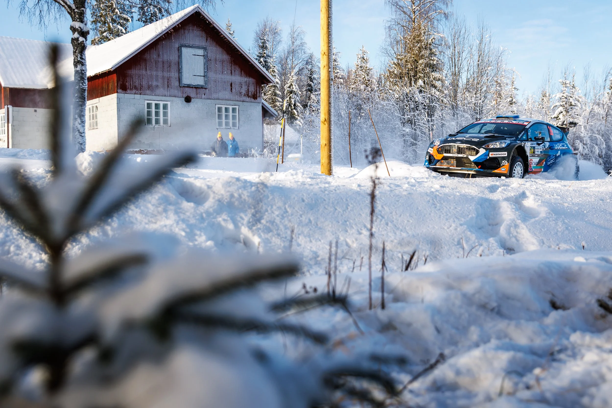 A rally car racing on a snowy winter landscape beside a rustic house with snowy trees in the background.