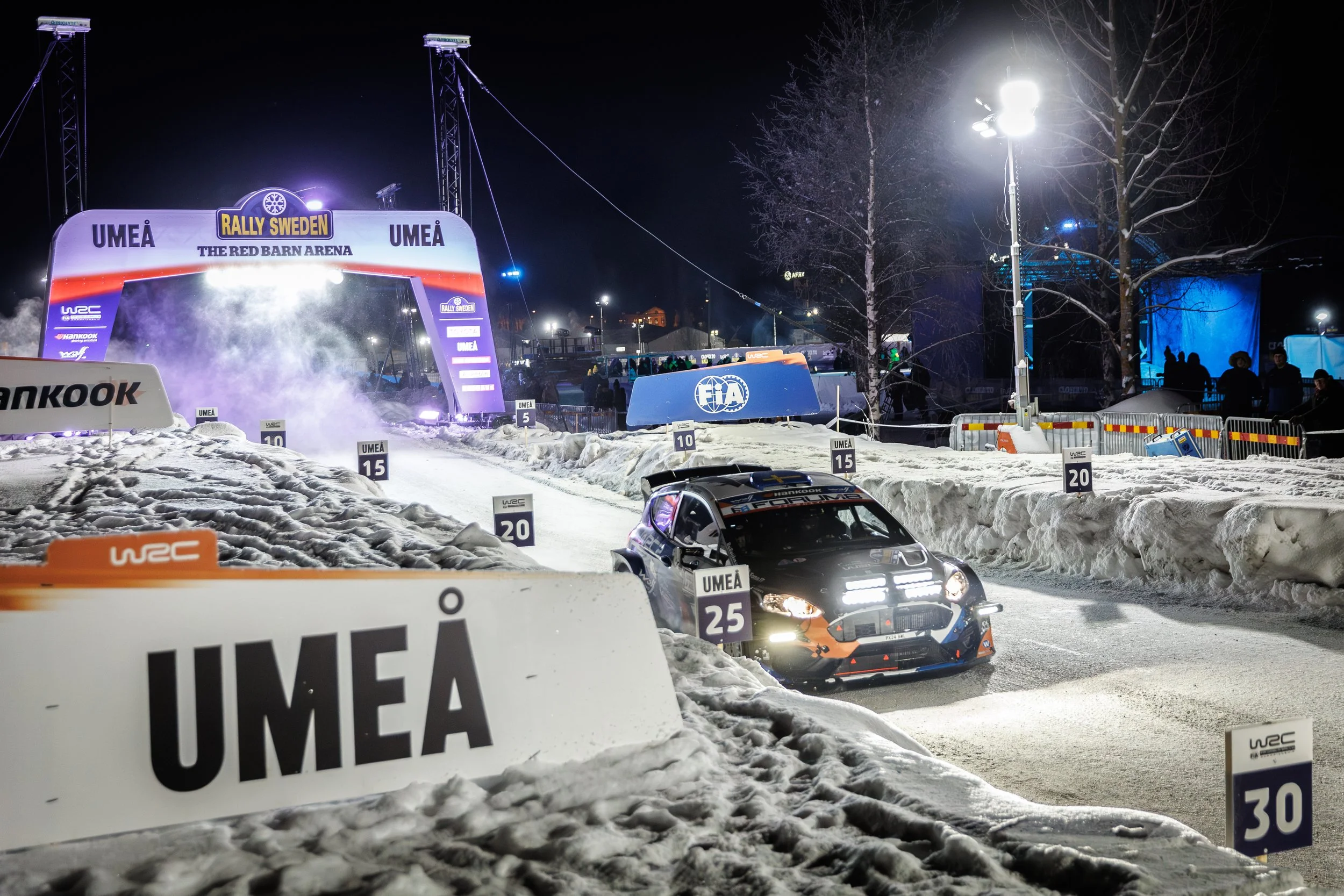 Nighttime scene of a rally race track covered in snow, with rally cars on the course and sponsor signs. The start/finish arch reads 'Rally Sweden' and 'Umeå,' with fog or smoke near the arch. Spectators stand behind barricades, and there are trees an