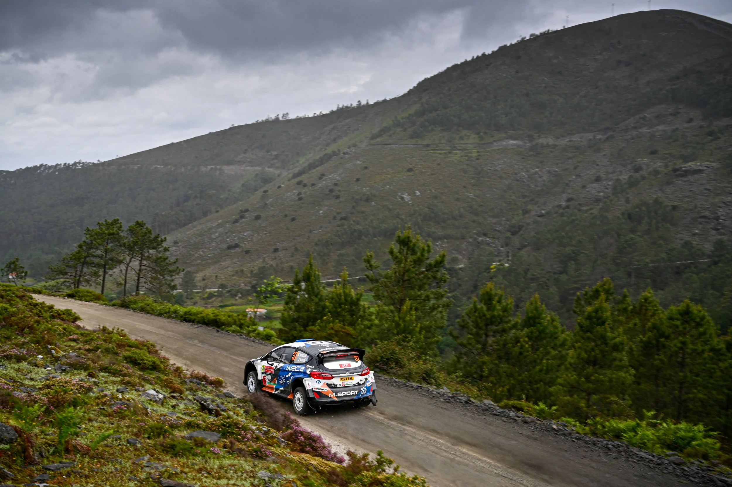 A rally car driving on a dirt mountain road with a scenic green landscape and cloudy sky in the background.