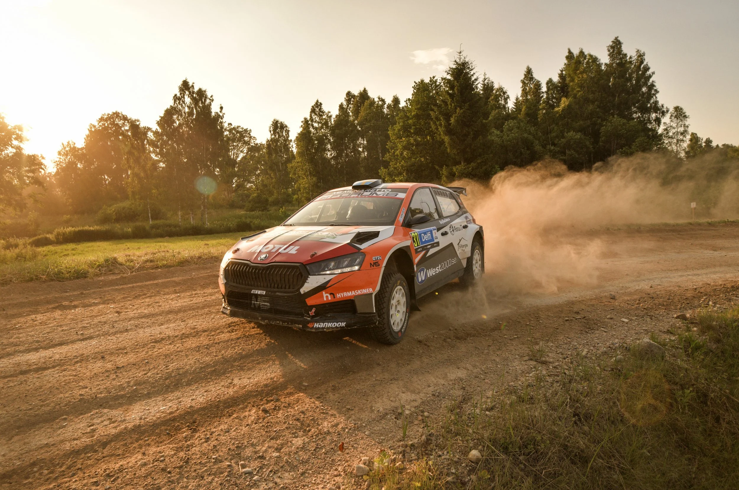 A rally car speeding along a dirt road with dust trailing behind, surrounded by trees during sunset.