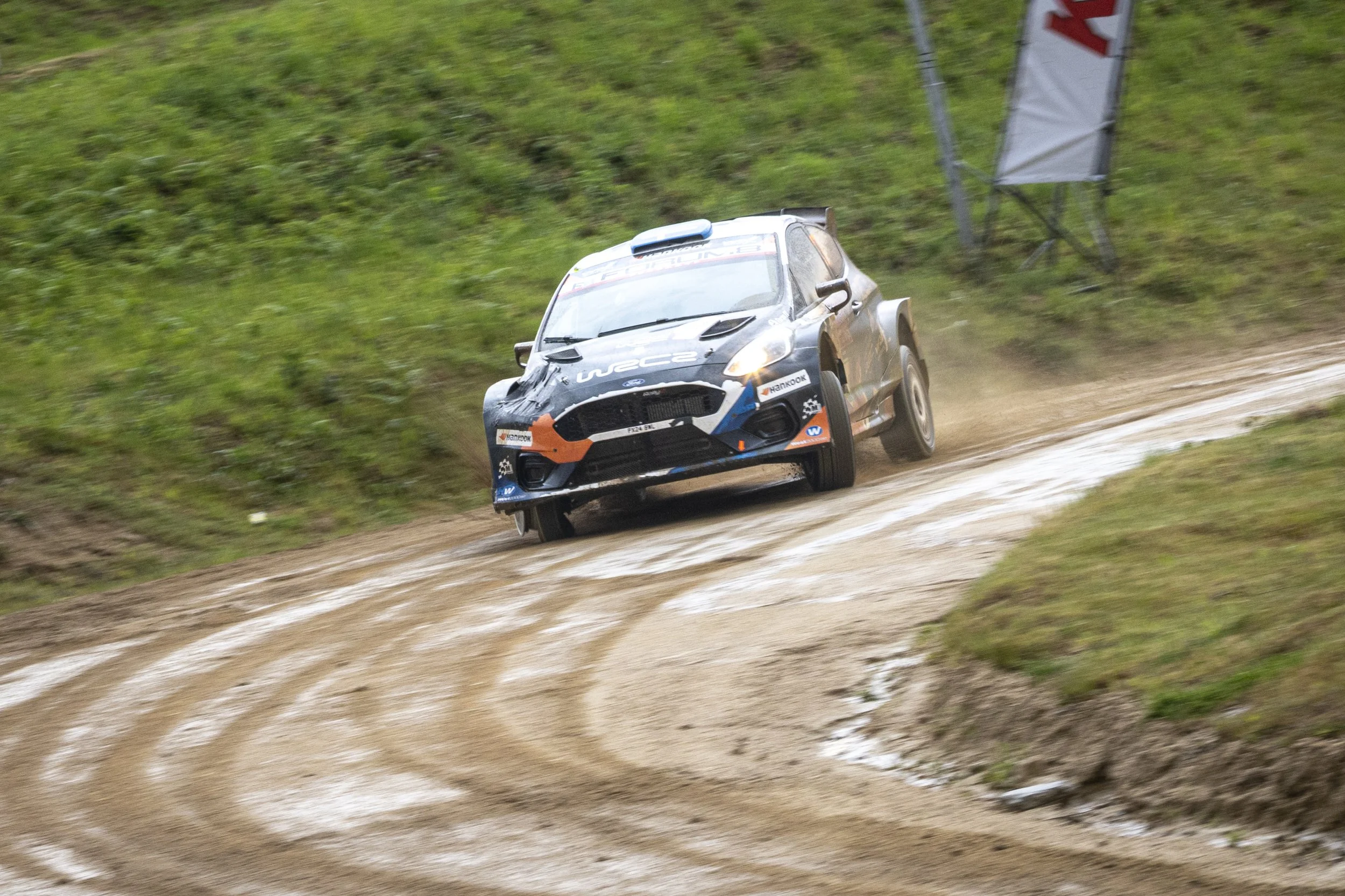 A rally car racing on a dirt track amid green grass and small bushes, kicking up dust behind it.