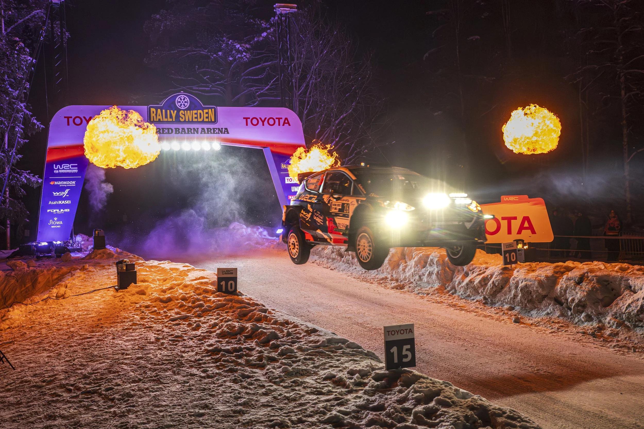 A rally car racing through snow at night during a competition, with flames and sparks flying from the vehicle, under an archway with banners, snow-covered ground, and trees in the background.