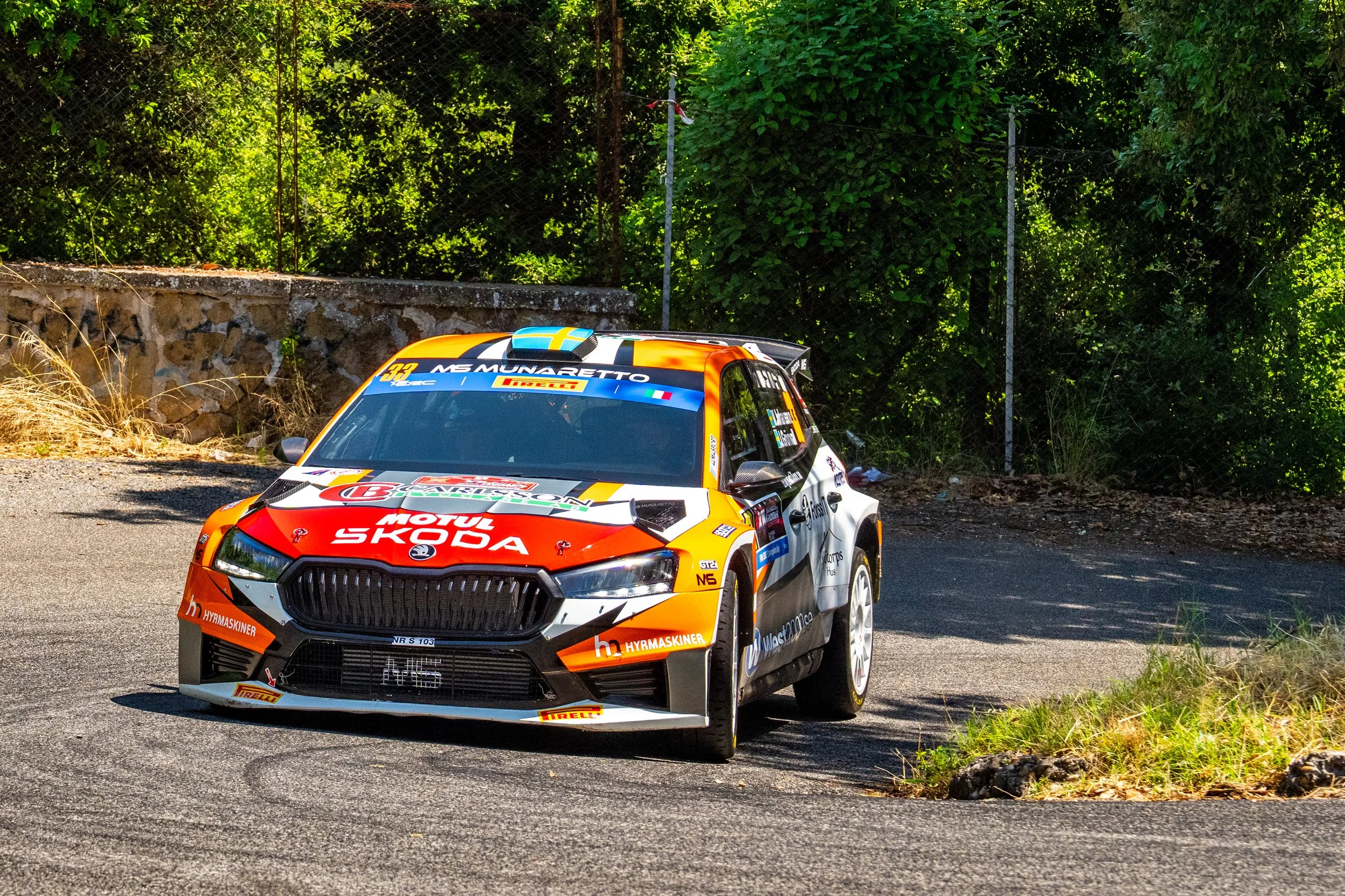 A rally car racing on a road surrounded by greenery, with a stone wall and wire fence in the background.