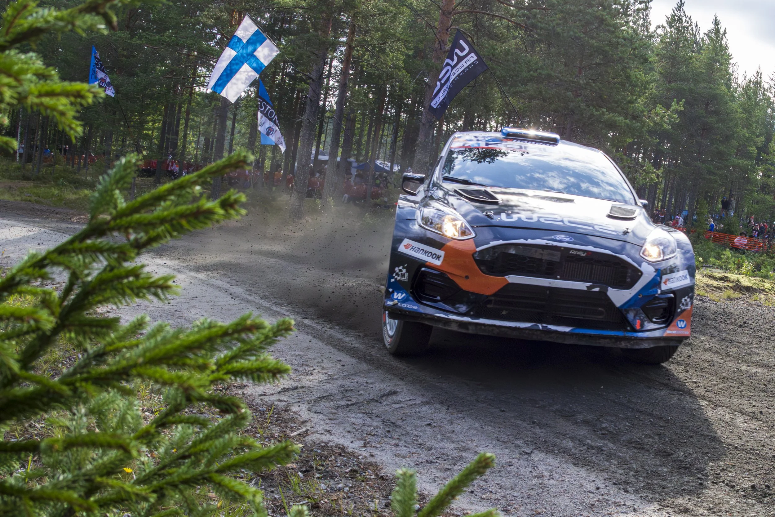 A rally car speeds around a corner on a dirt track in a wooded area, kicking up dust, with spectators and flags visible in the background.