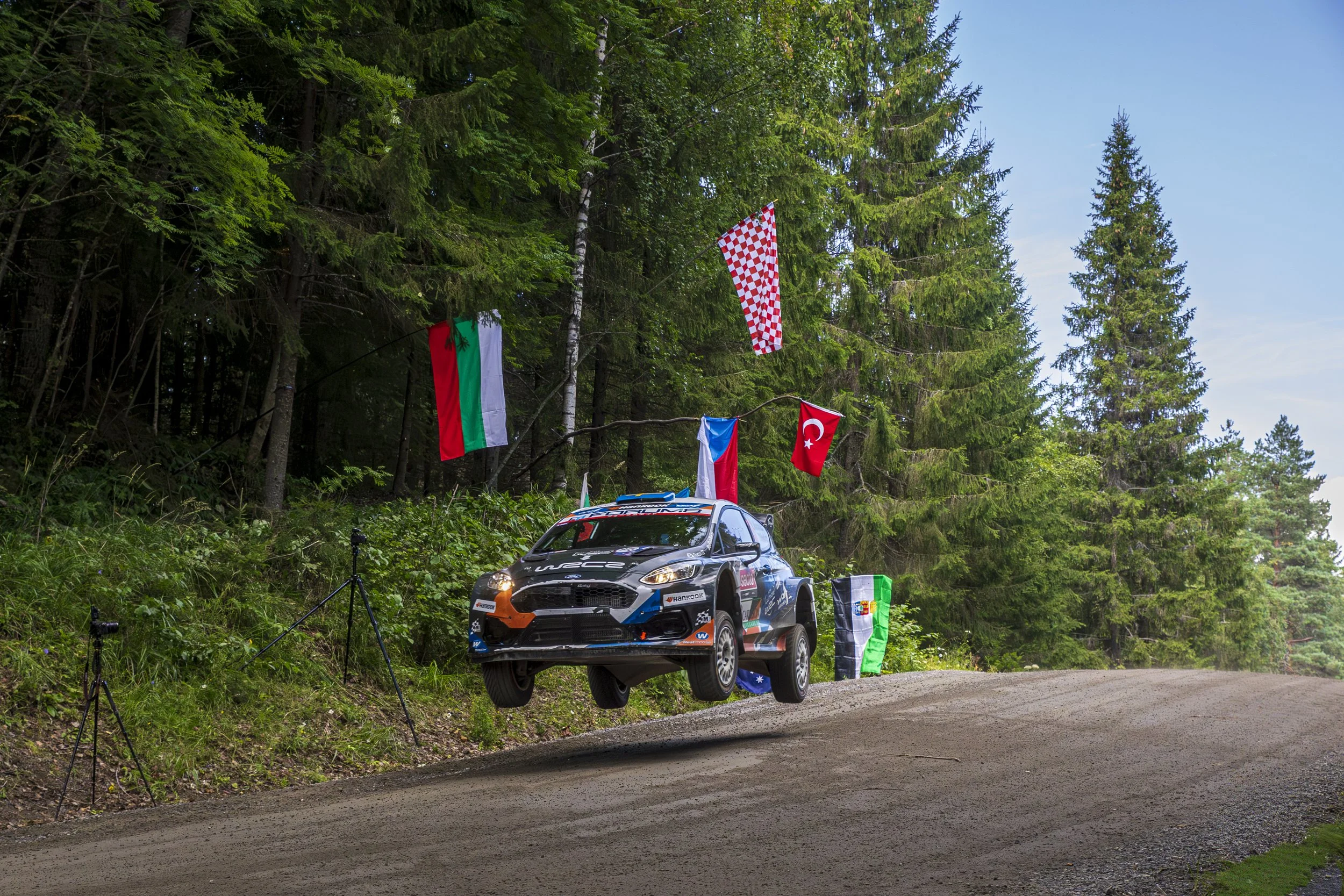 A rally car is mid-air, jumping off a dirt road during a race, with green forest and flags of various countries, including Bulgaria, Croatia, Turkey, and Australia, hanging from trees in the background.