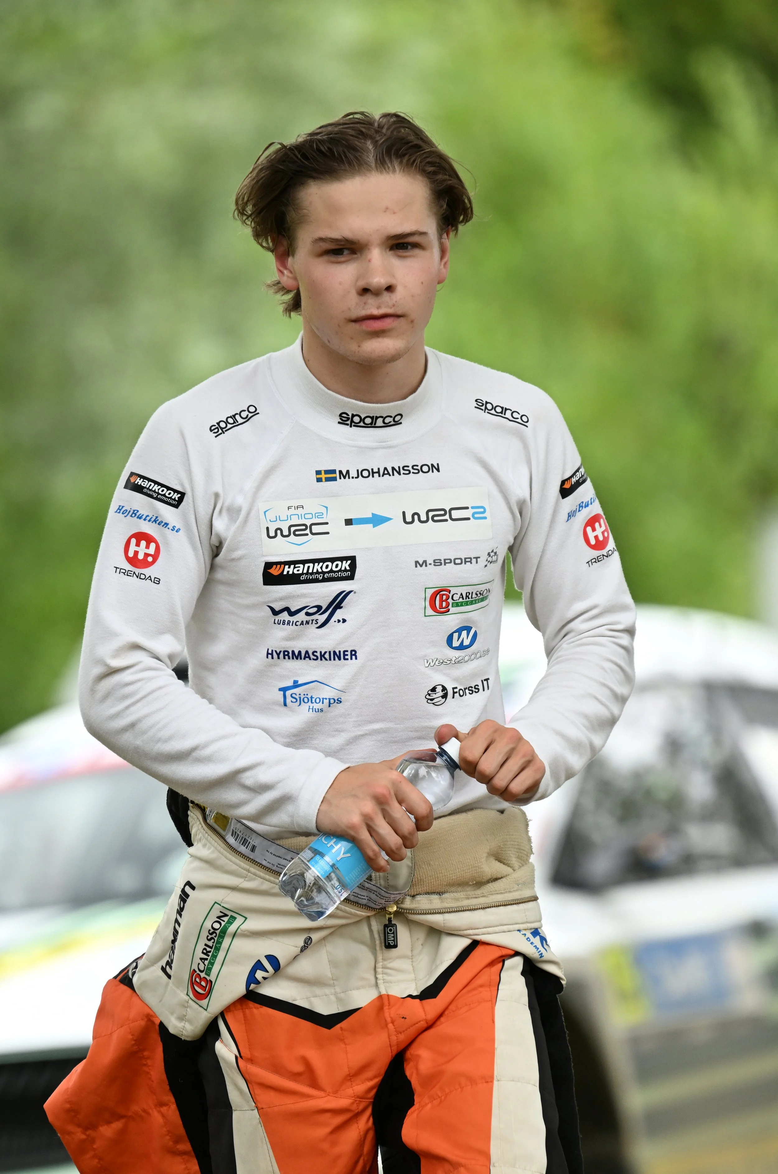 Young male race car driver wearing a white racing suit with multiple sponsor logos, holding a water bottle in front of a blurred green outdoor background.