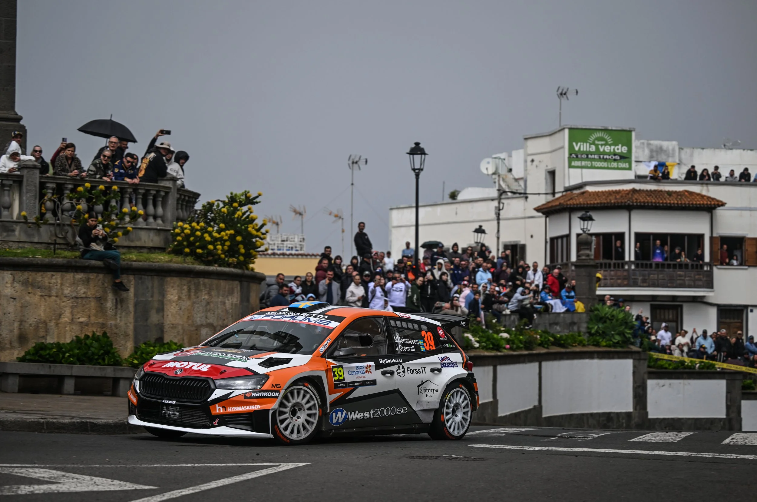 A rally car on a city street with a crowd of spectators on a raised platform and sidewalk, overcast sky, buildings, and streetlights in the background.