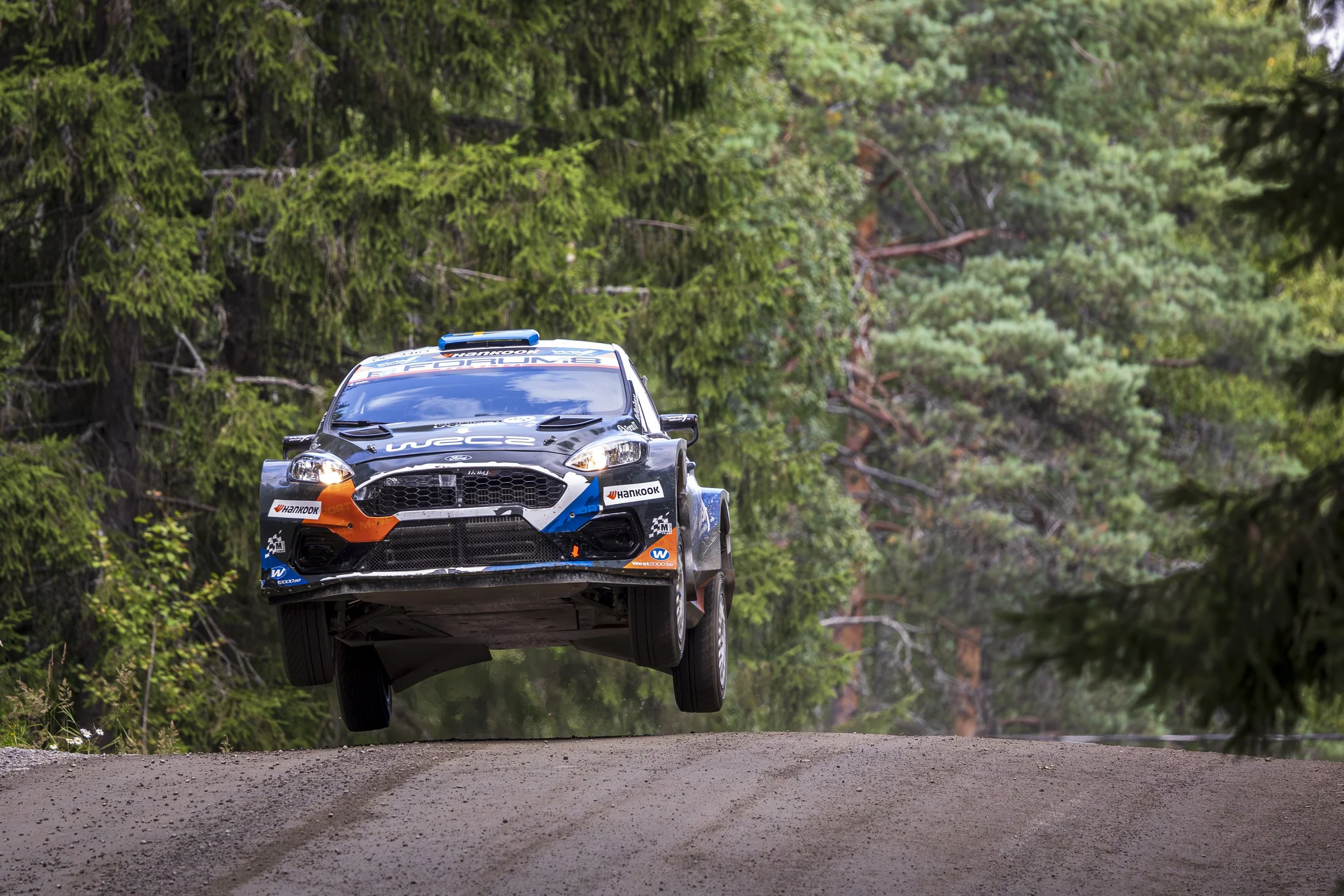 Rally car airborne above dirt road surrounded by green trees.