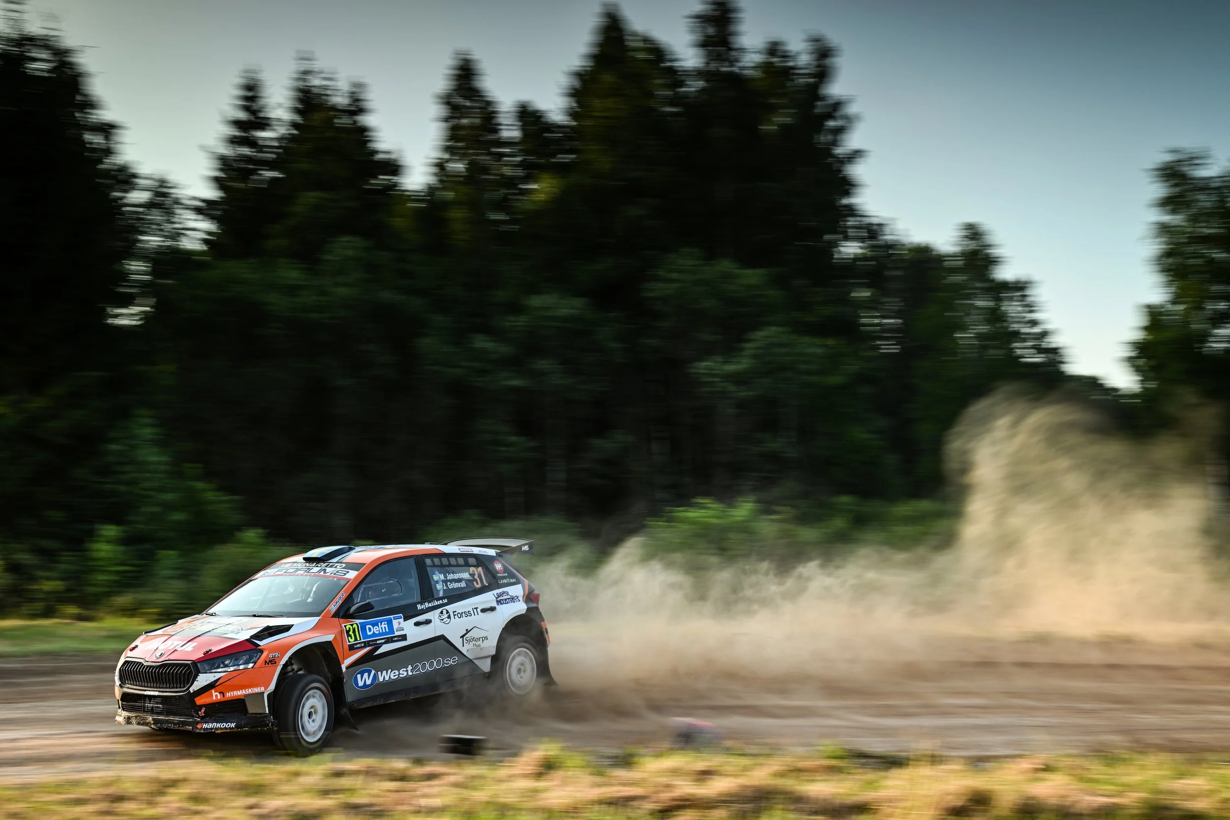 Rally car speeding on dirt road in forest, kicking up dust, during a race.