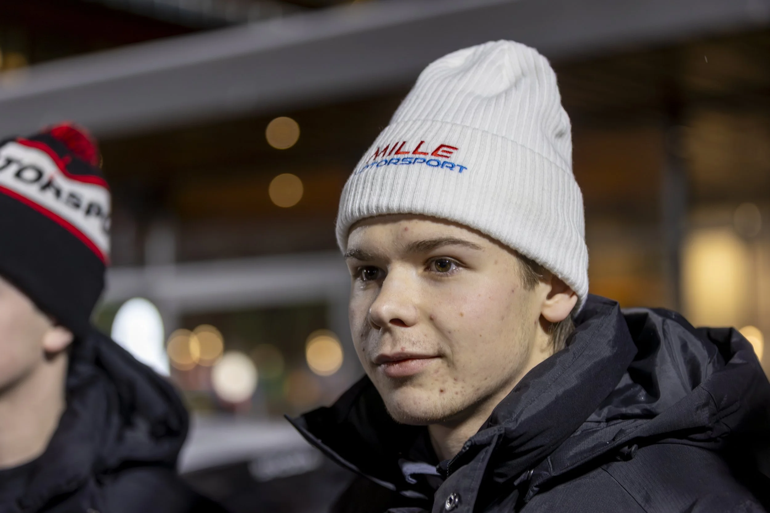 A young man in a white knit beanie with red and blue text, wearing a dark jacket, looking intently at another person during nighttime outdoors.
