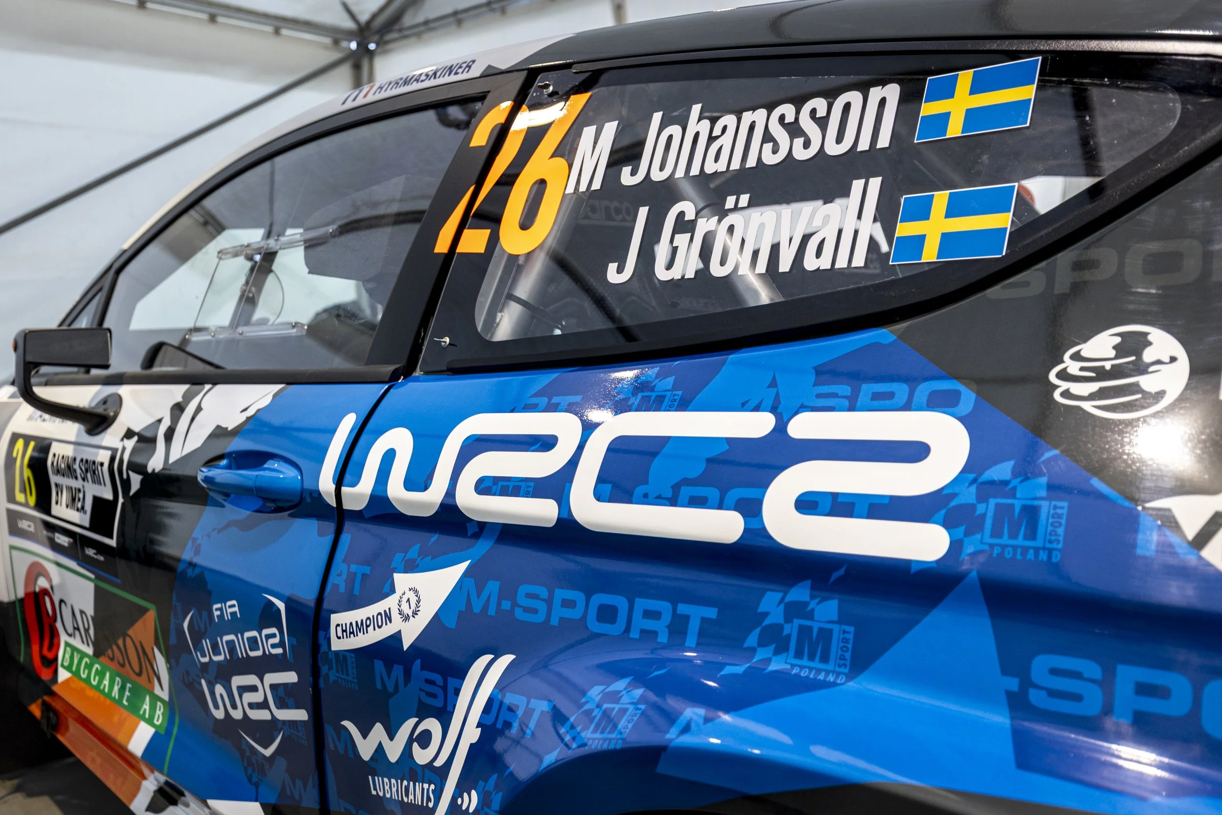 Close-up of a rally car with sponsor logos, driver names Johansson and Grönvall, and Swedish flags on the window.