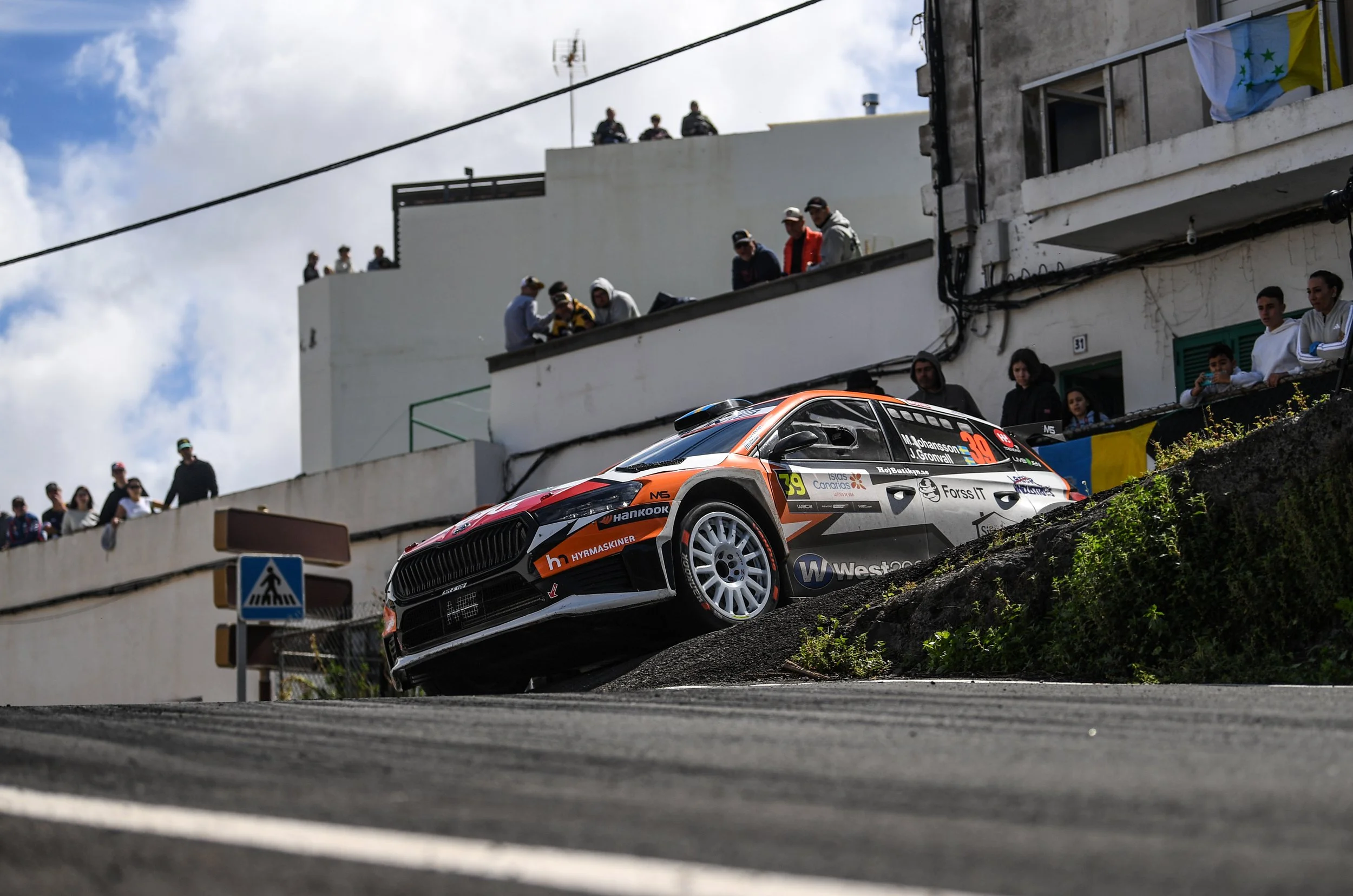 A rally car, number 39, is racing downhill on a dirt track, with spectators standing on a terrace above.