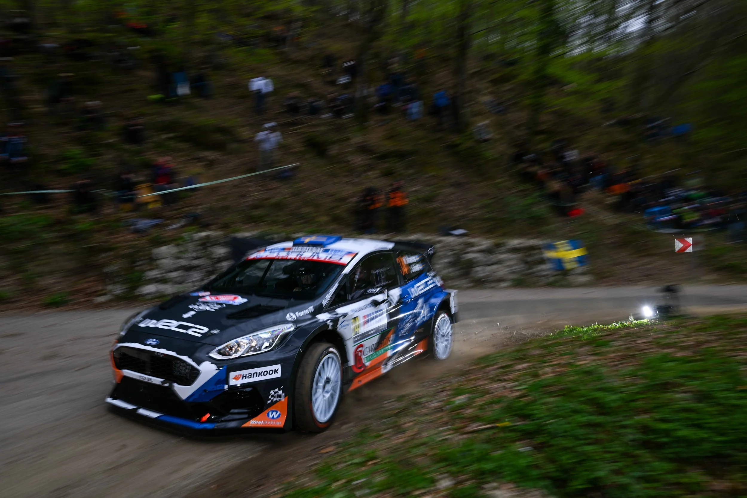 A rally car speeding on a dirt road with a forested hillside in the background, spectators watching from the hillside.