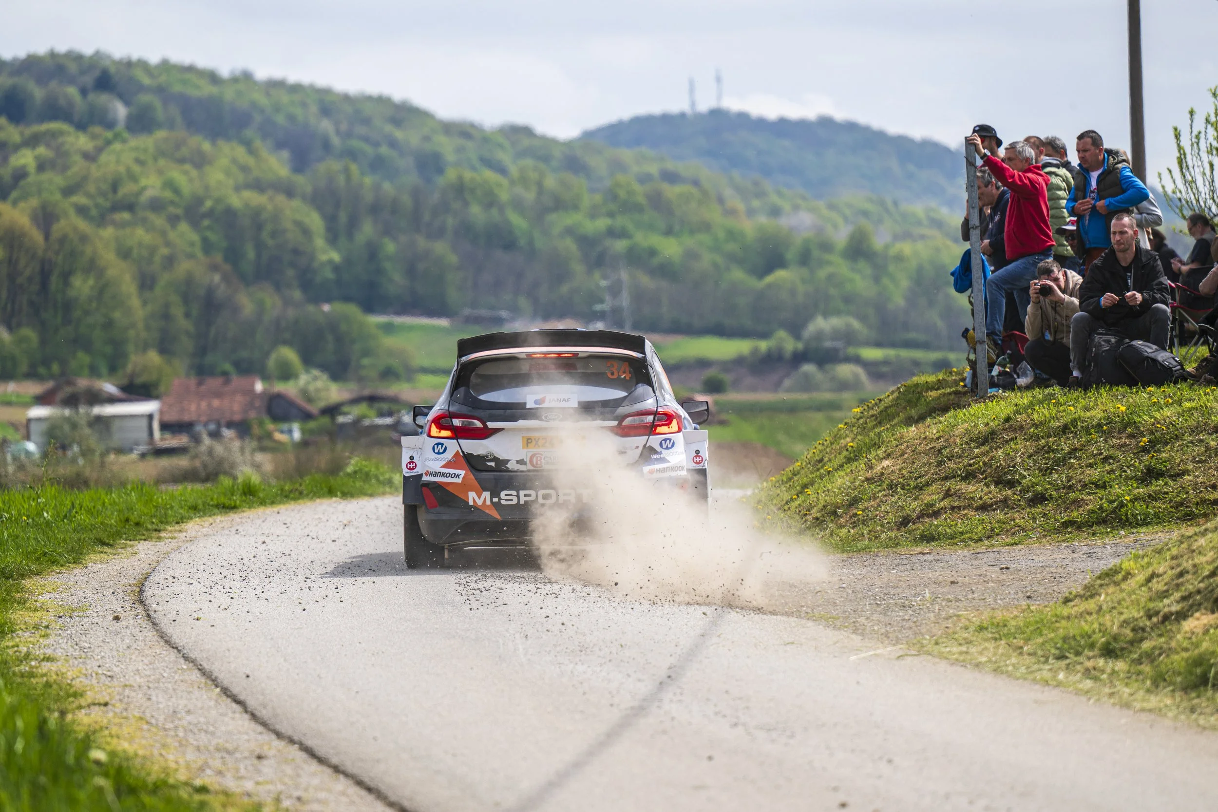 A rally car with sponsor logos speeds down a gravel road, kicking up dust, with photographers on the right side taking pictures of the race, and a scenic, hilly landscape in the background.