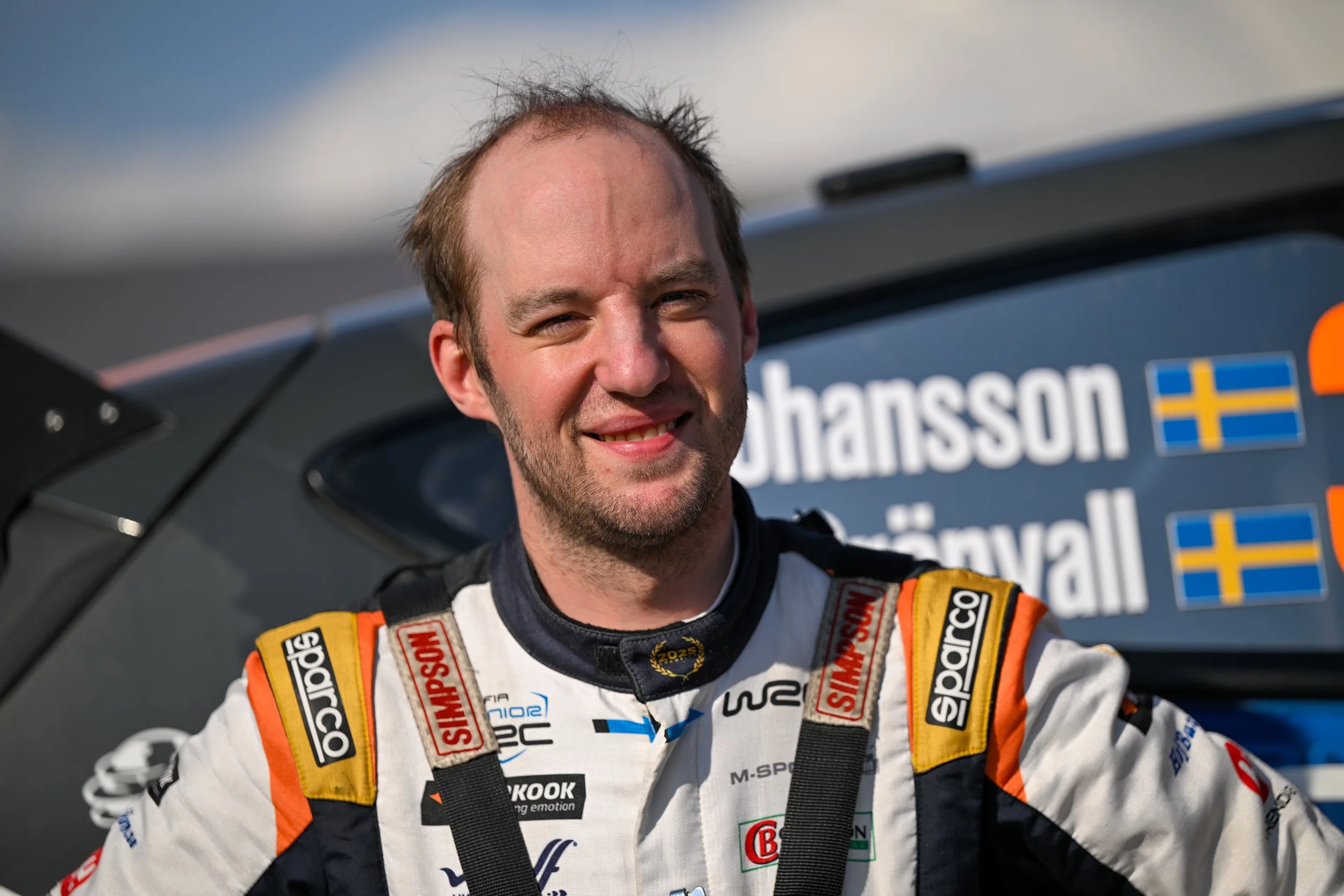 Smiling man in racing suit standing in front of race car with sponsor and team logos.