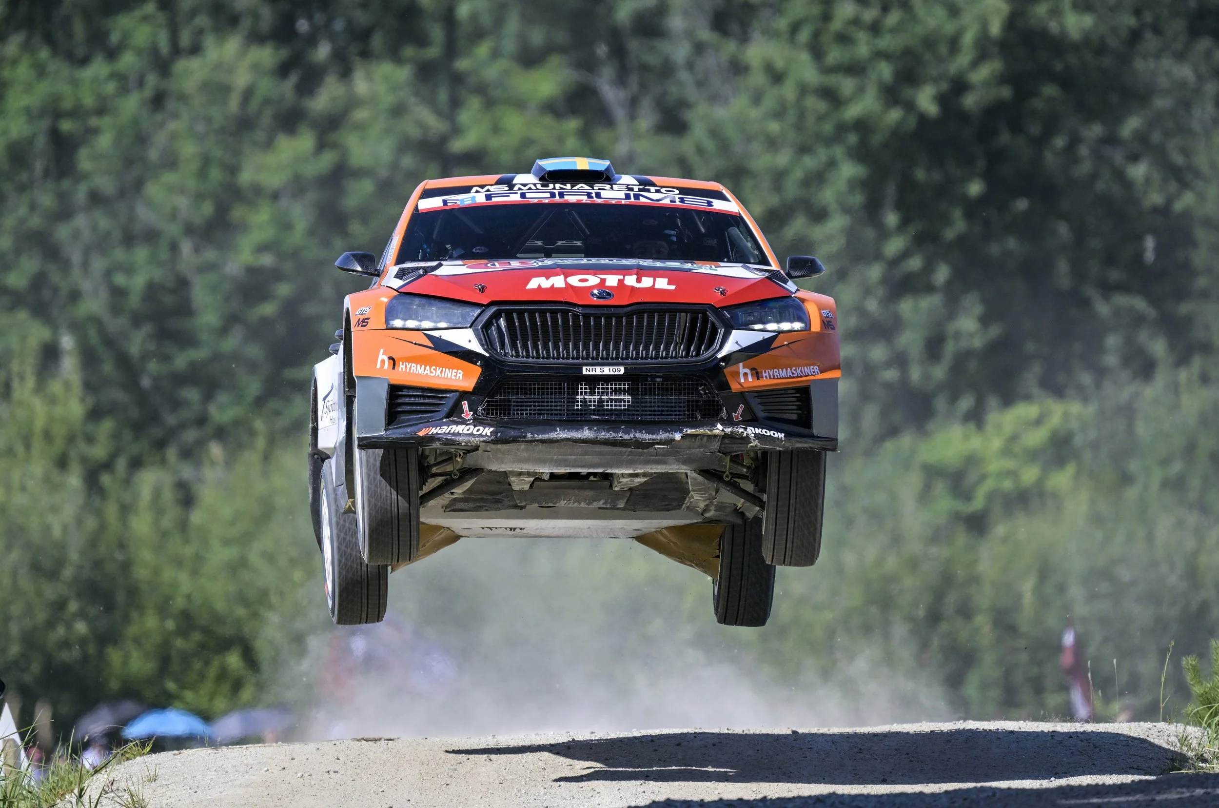 A race car flying through the air during a rally, with a dust cloud trailing underneath, set against a background of trees and greenery.