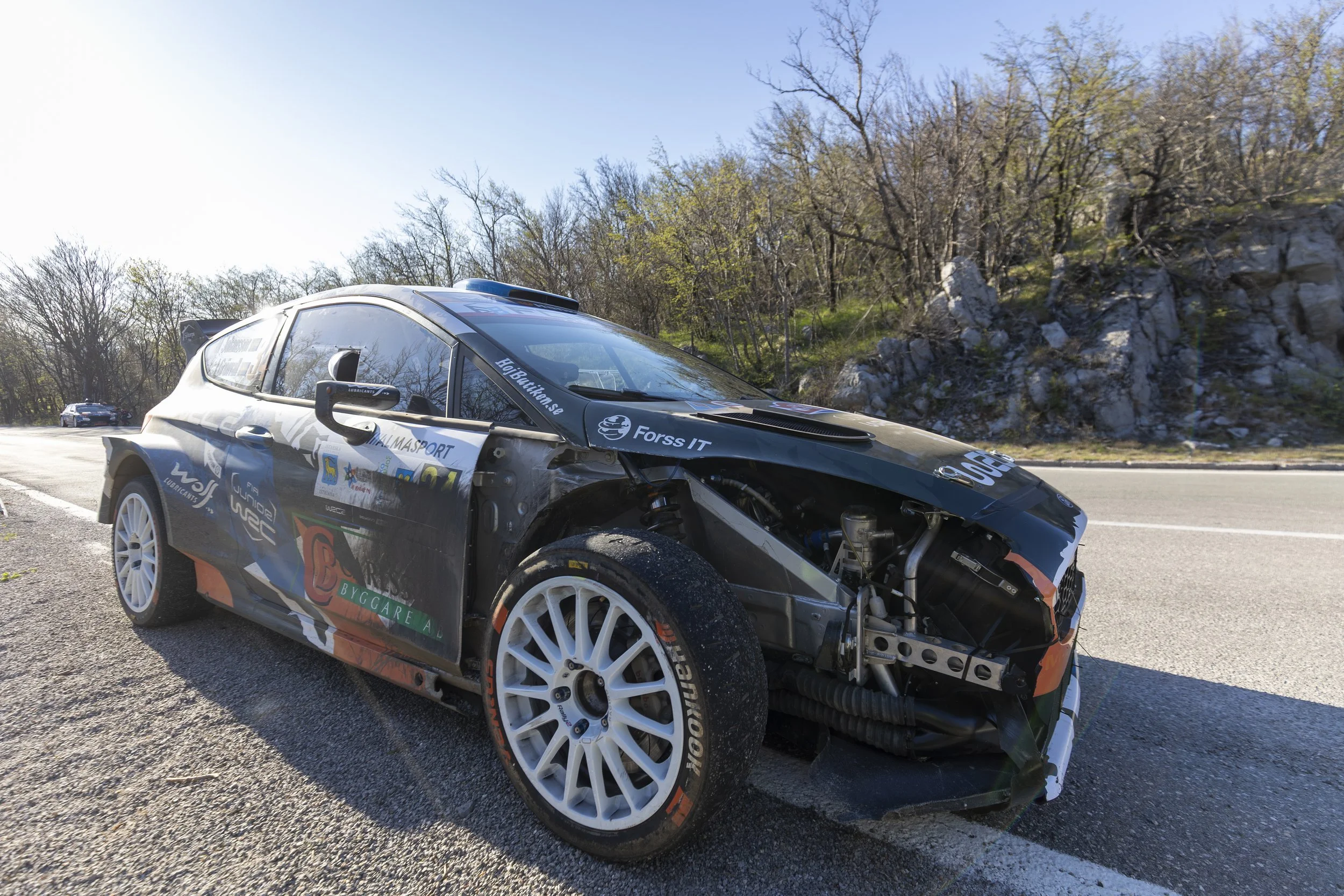 Damaged rally car with missing front bumper and exposed mechanical parts on a roadside with trees and rocky hillside in the background.
