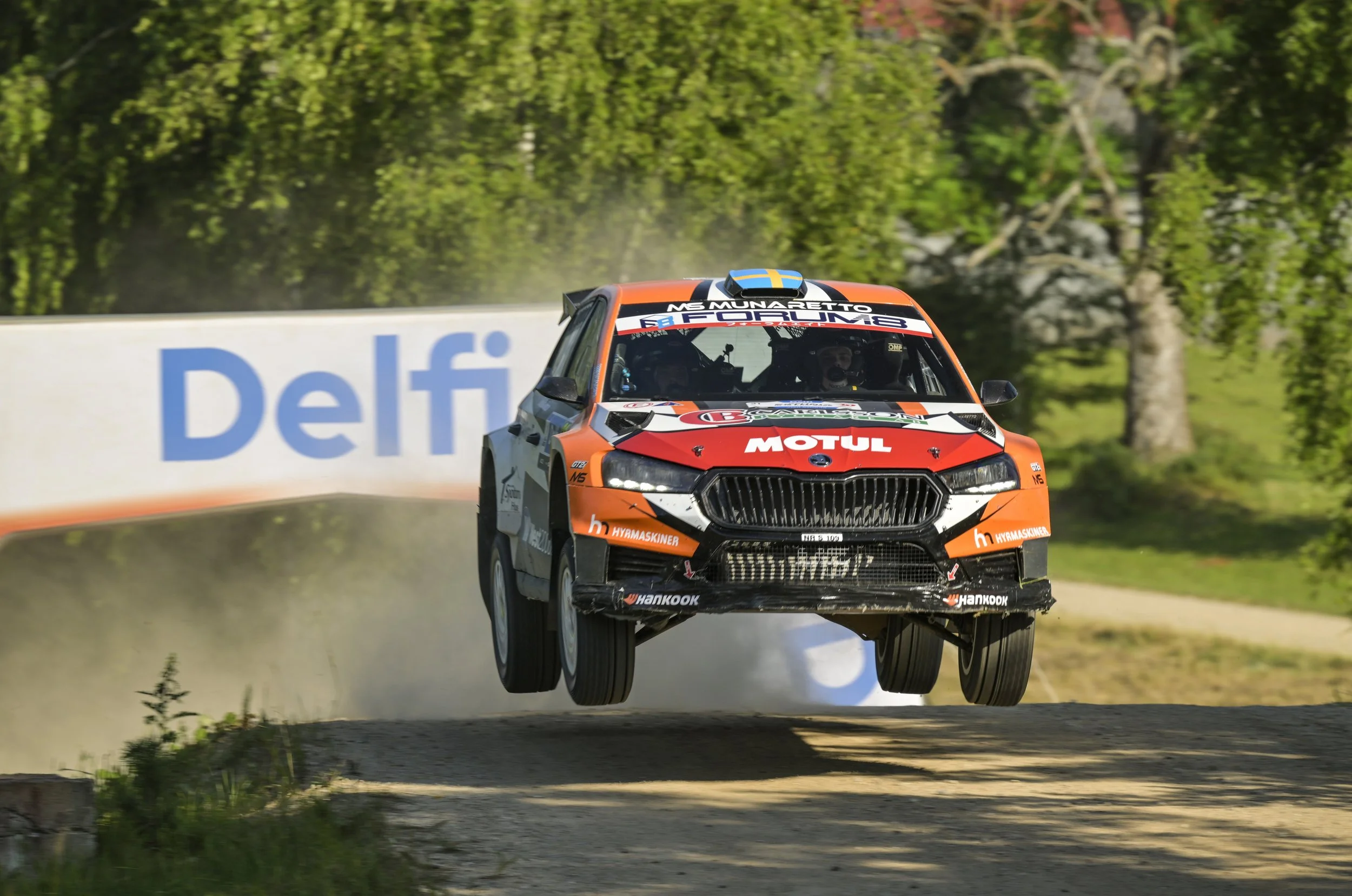 A rally car airborne over a dirt road with a dust cloud behind it, surrounded by green trees, during a race.