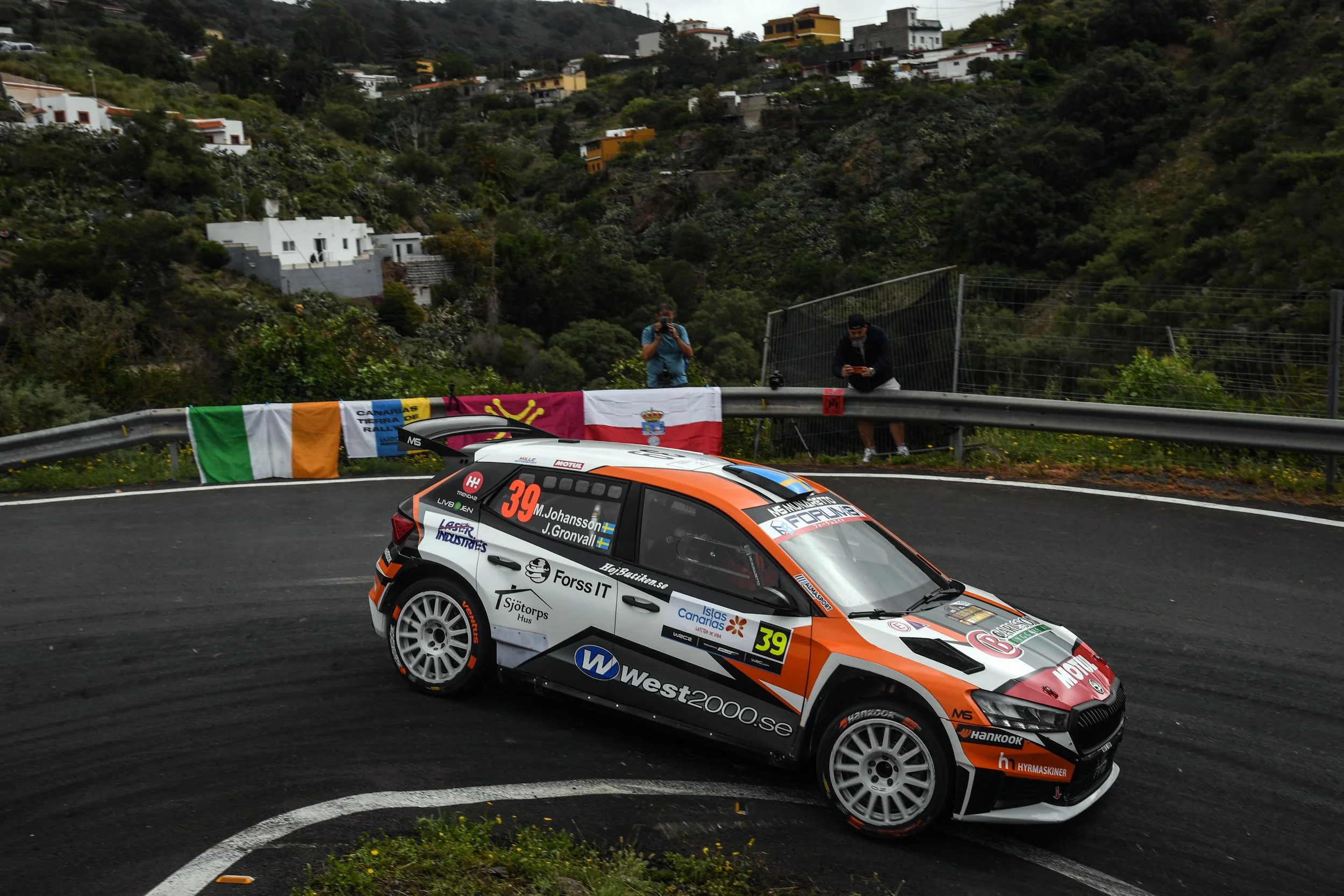 A rally car with number 39 racing on a winding road with a hilly residential area in the background and two people taking photos beside the road.