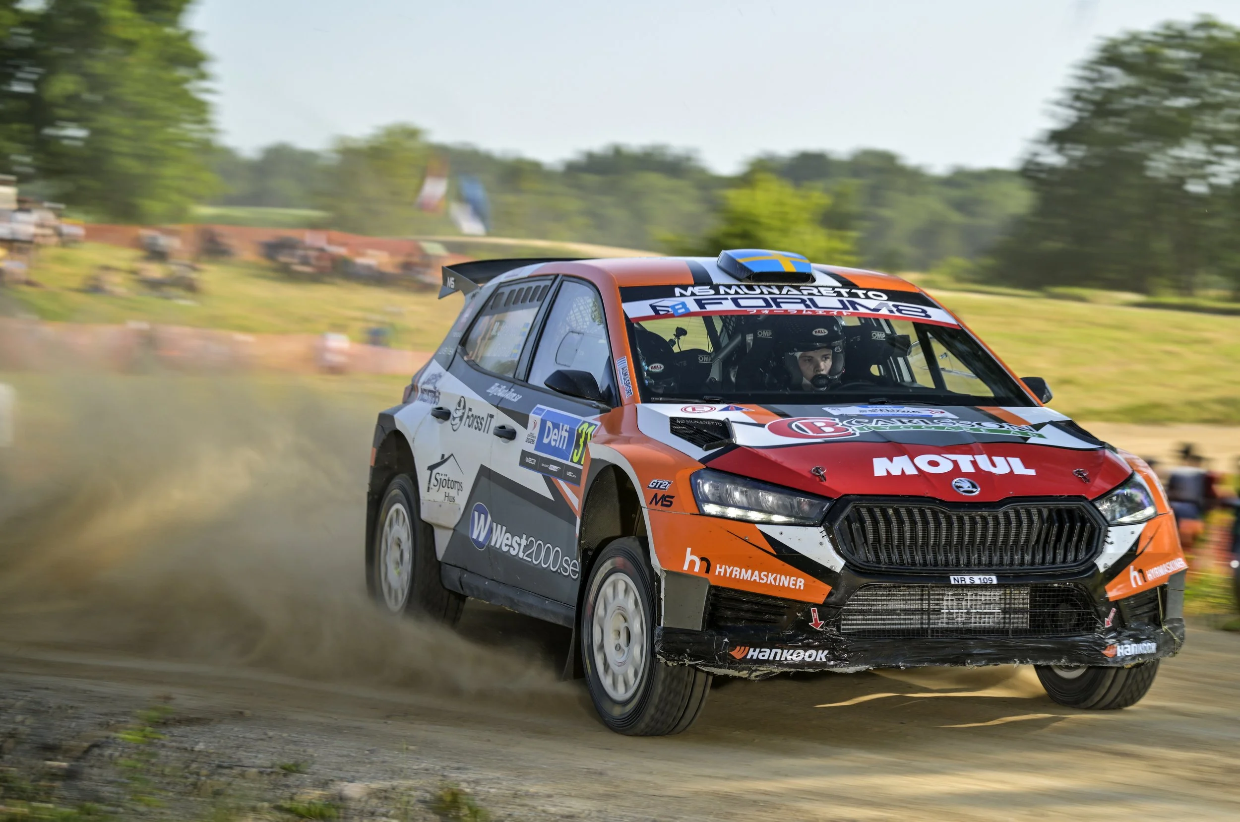 A rally car speeding on a dirt track during a race, with trees and spectators blurred in the background.