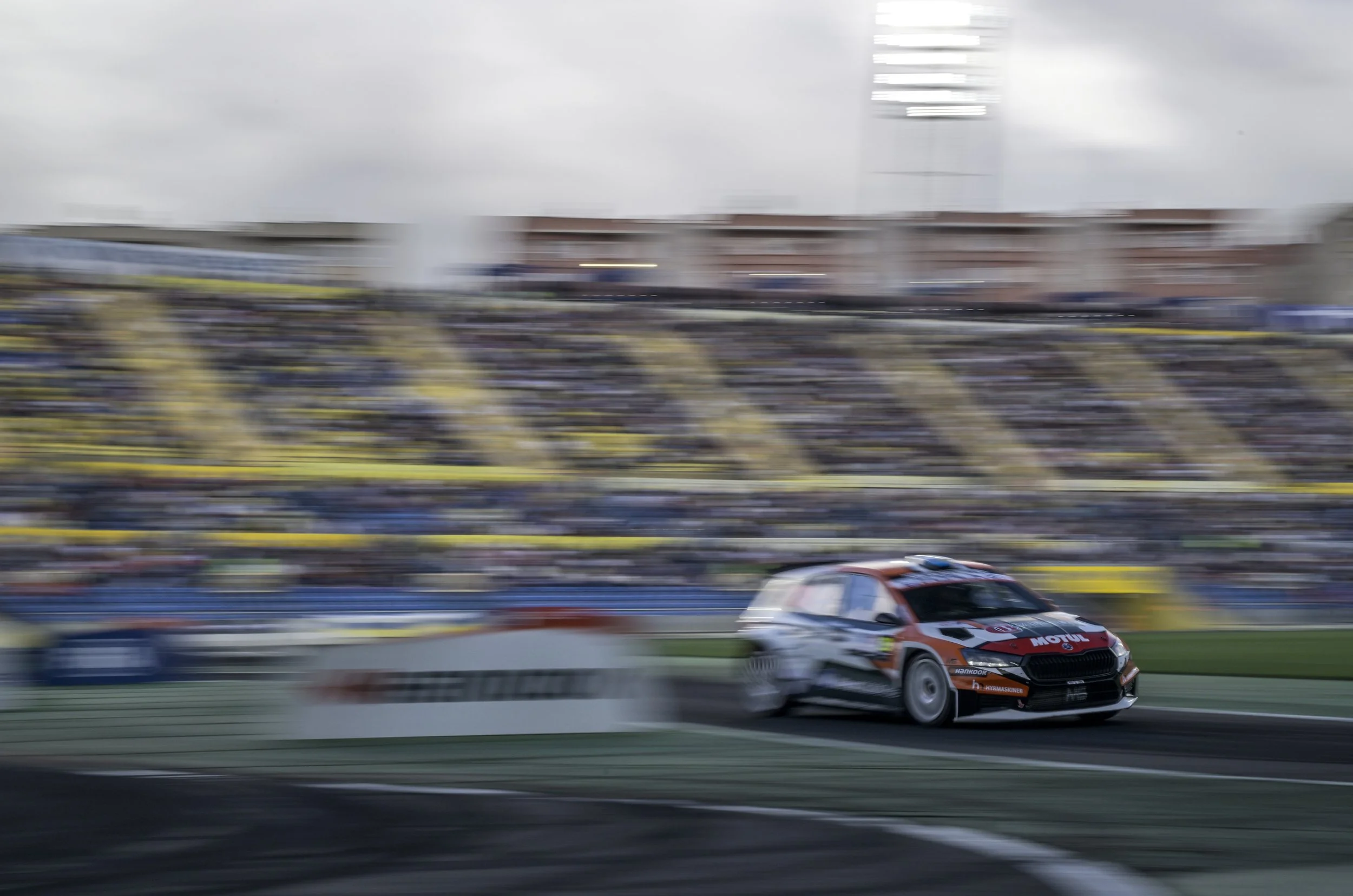 A race car on a racetrack with a blurred background showing stadium seating and a cloudy sky.