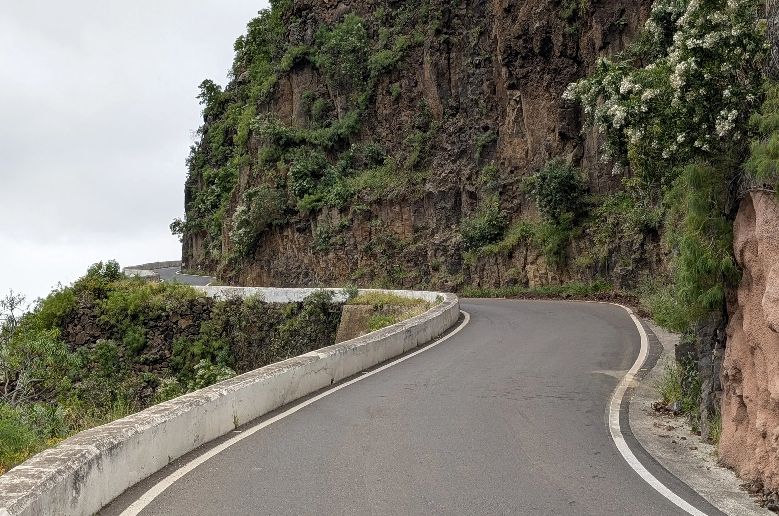 A winding mountain road with a rock wall on one side and a cliff with green vegetation on the other, under an overcast sky.