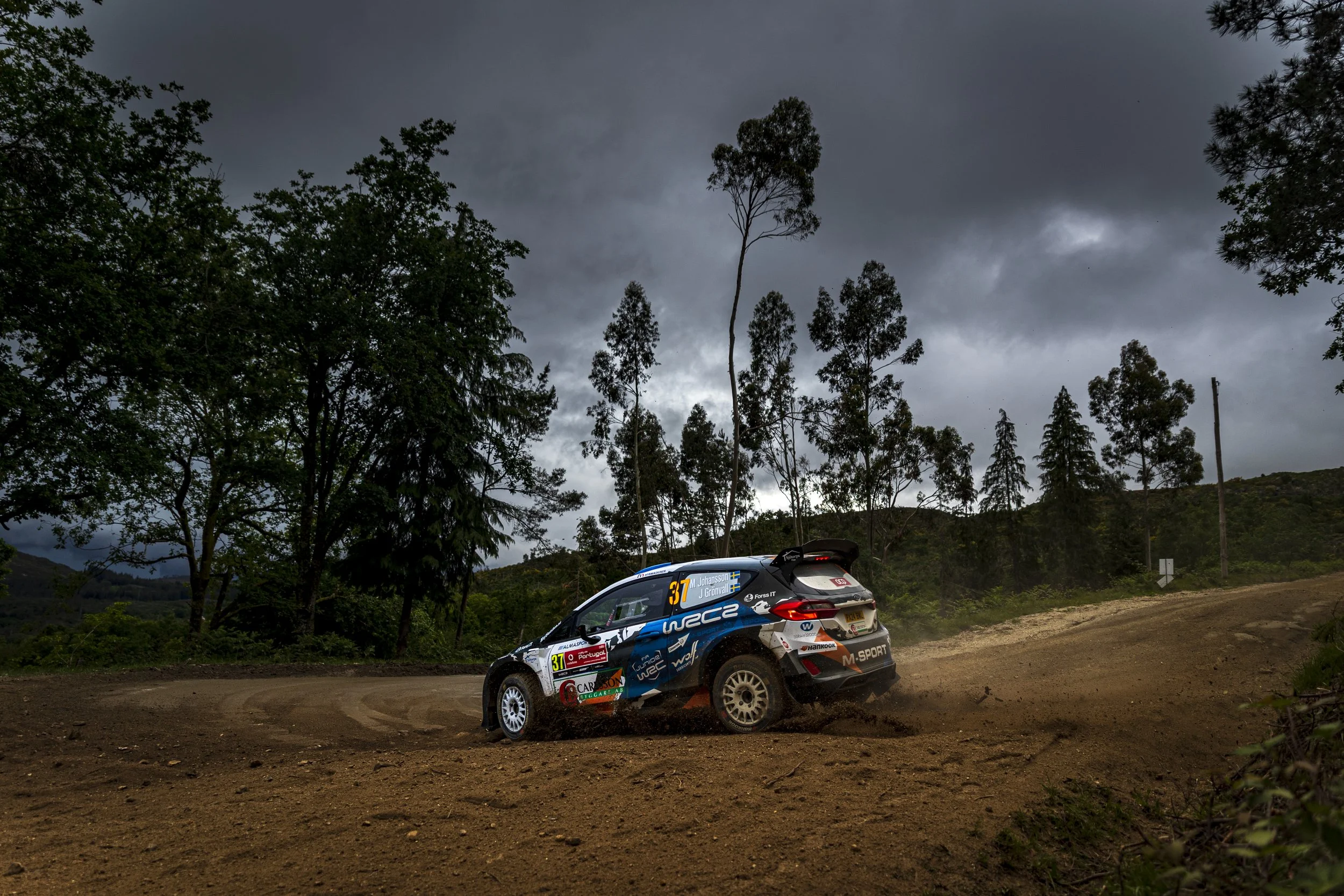 A rally car speeding on a dirt road outdoors with trees and dark cloudy sky in the background.