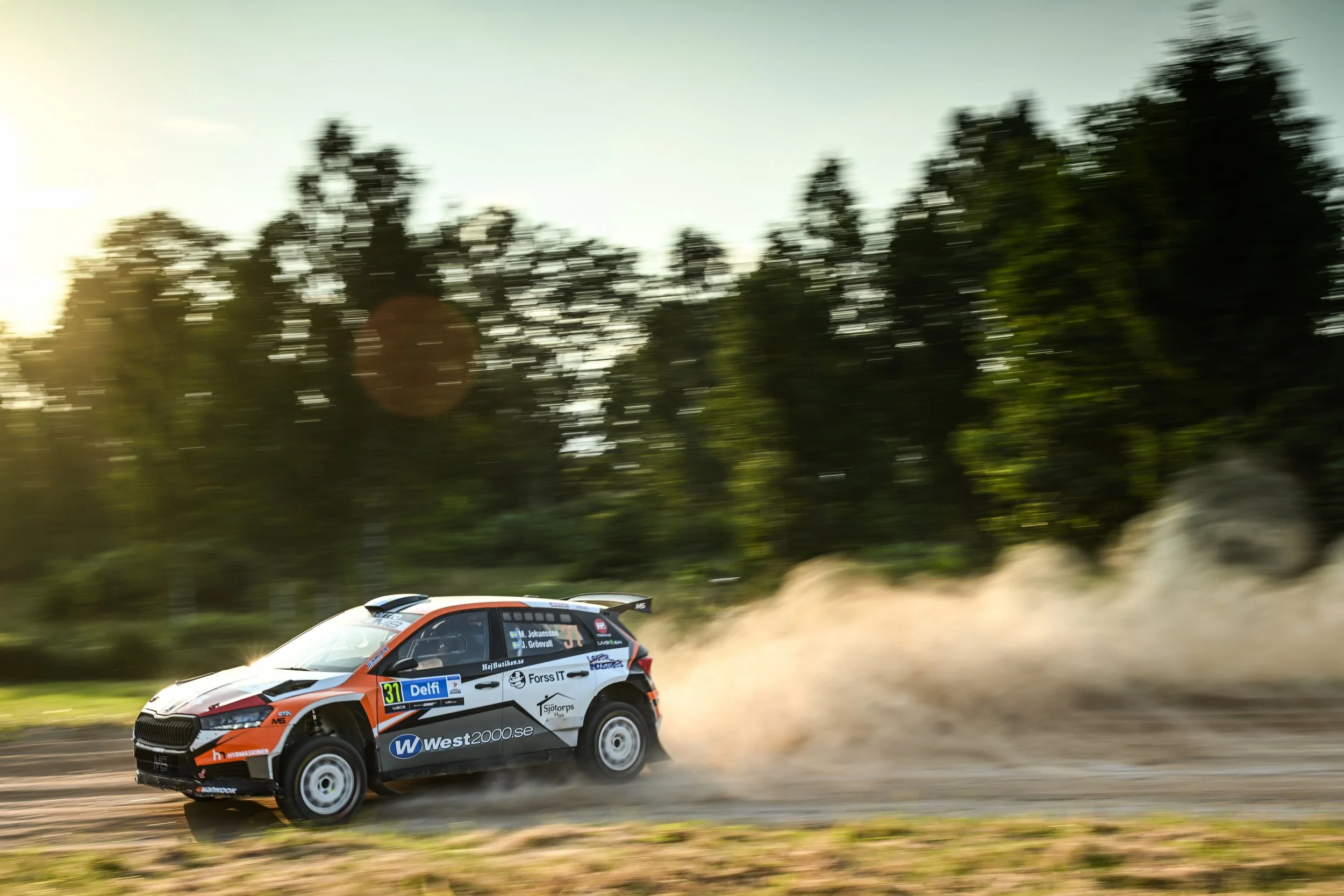 A rally car racing on a dirt track with trees and dust clouds in the background, during a daytime event.