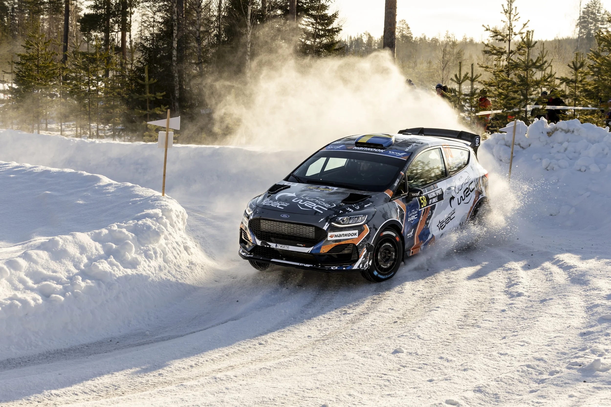 A rally car race through a snowy forest, kicking up snow dust.