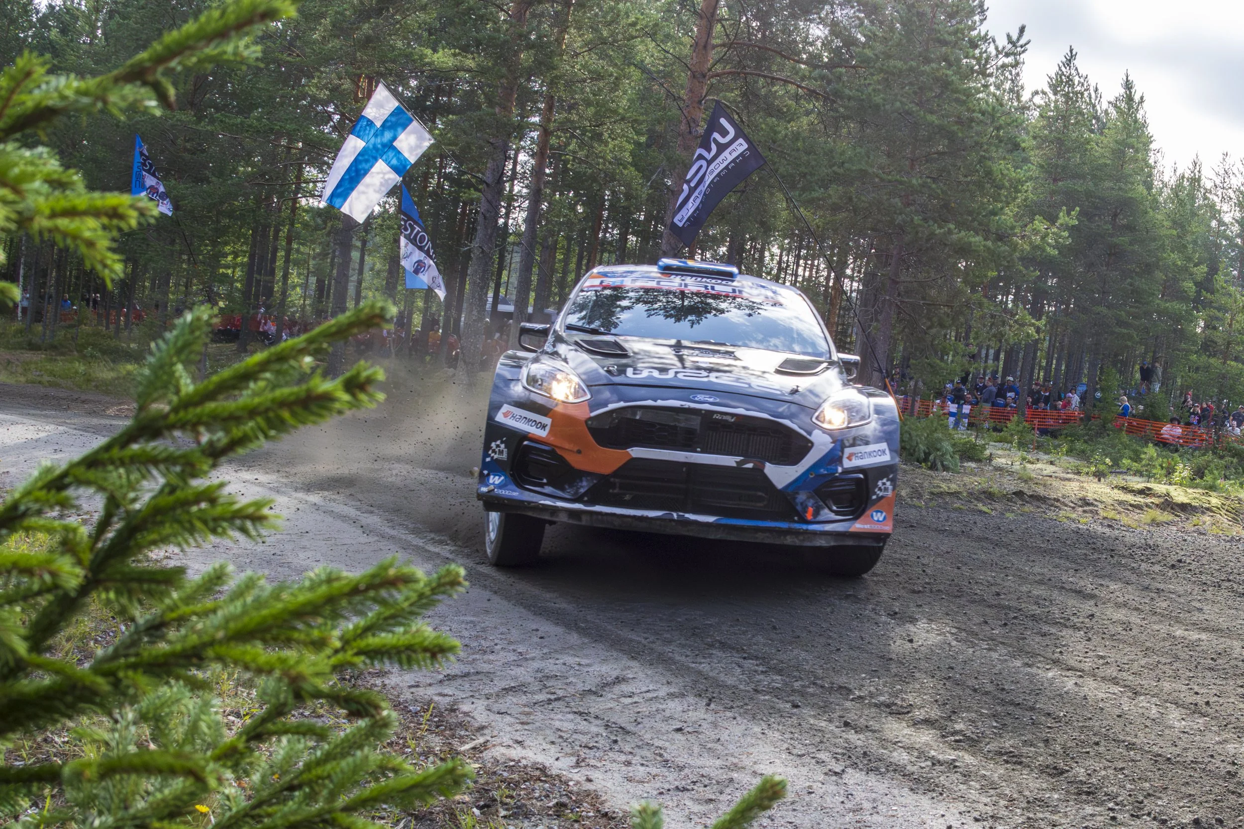 A rally car racing on a dirt forest track, with flags of Finland and other race banners in the background, surrounded by trees and spectators.