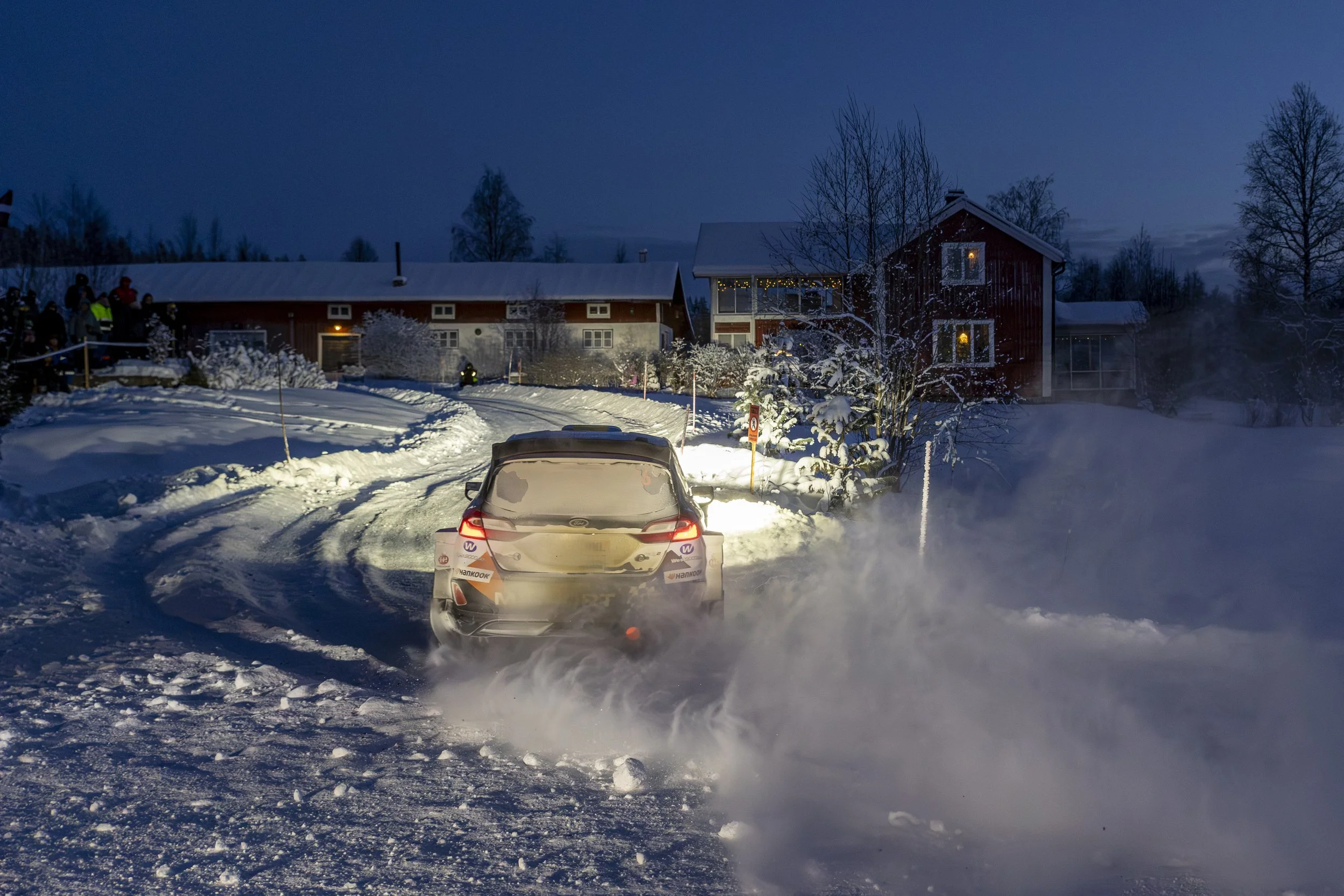 A car driving through snow at night, kicking up snow behind it, with a house illuminated by warm lights in the background.