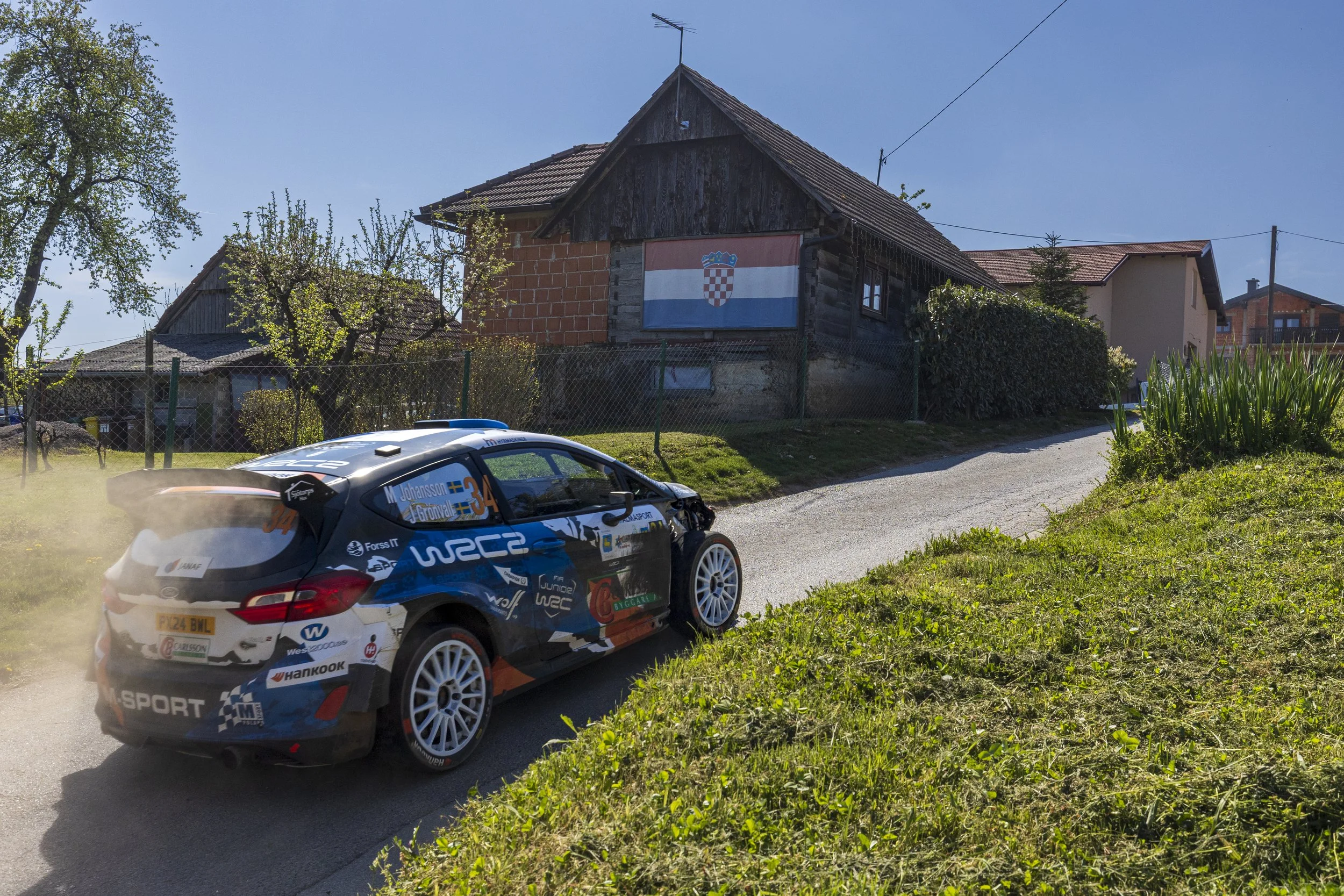 A rally car racing on a dirt road past a rustic house with a Croatian flag hanging on the wall. The car is decorated with various sponsor logos and the drivers' names.