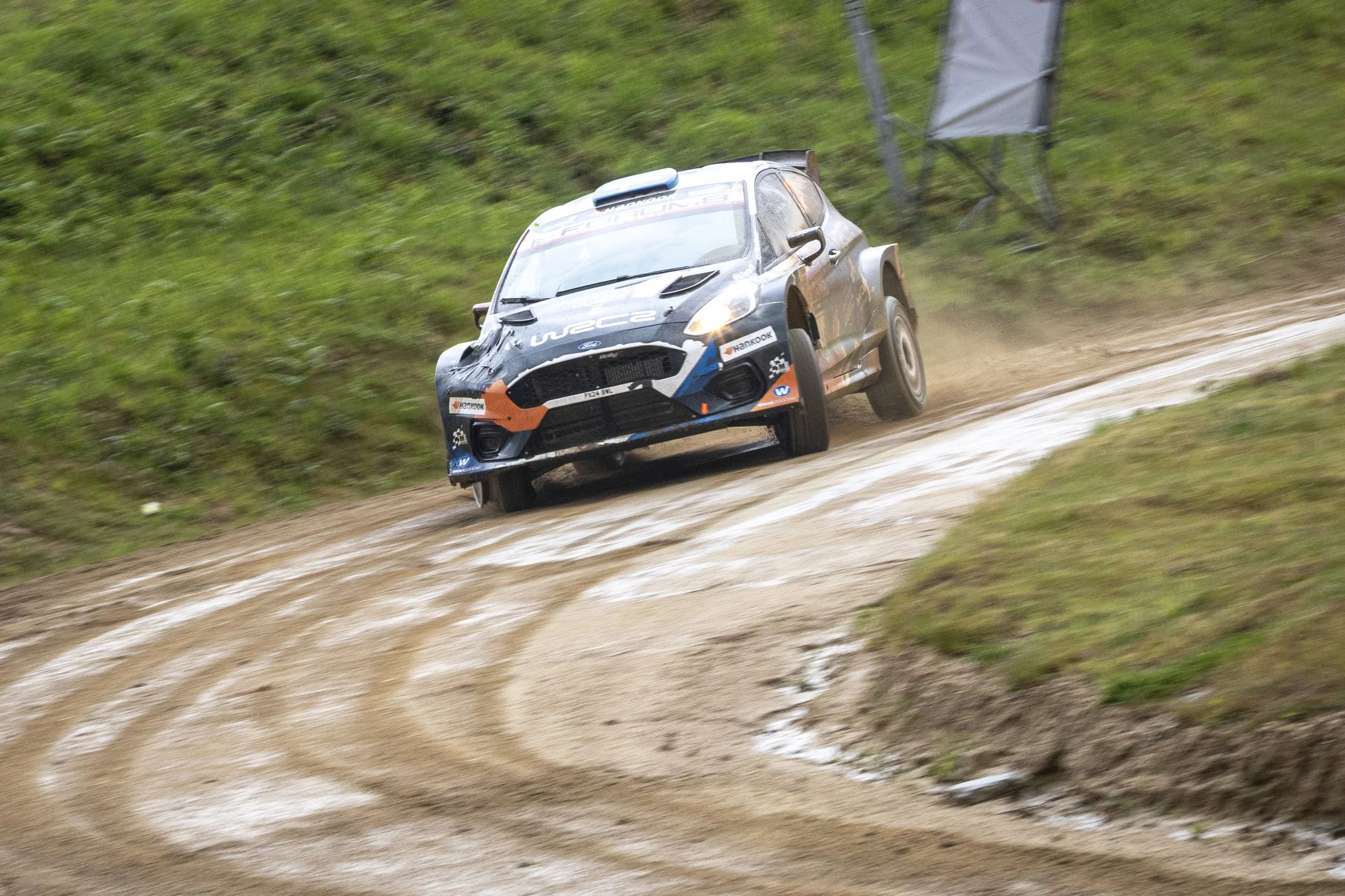 A rally car speeding on a dirt road with grass on either side, kicking up dust behind it.