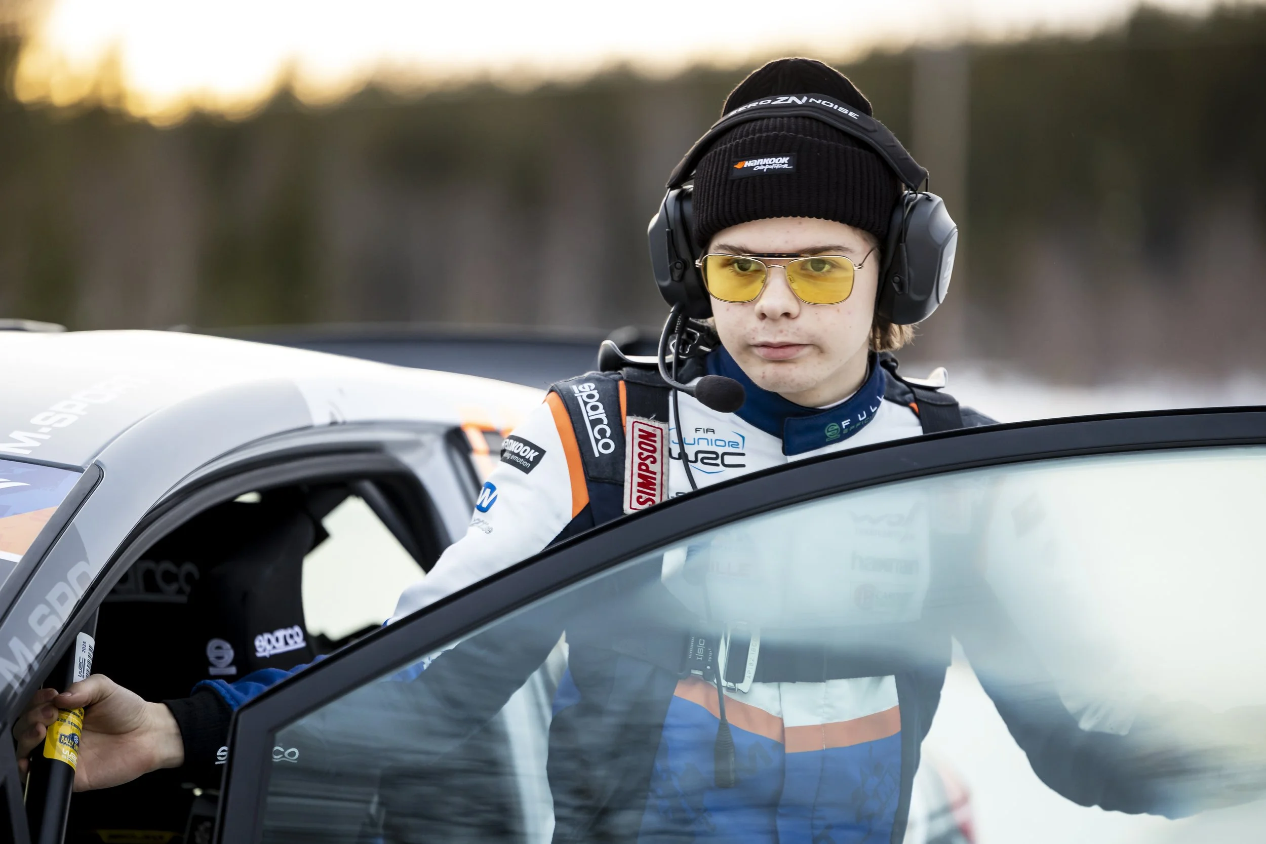 A young race car driver with blond hair, wearing yellow-tinted glasses, a headset, and a racing suit, is standing outside a car.