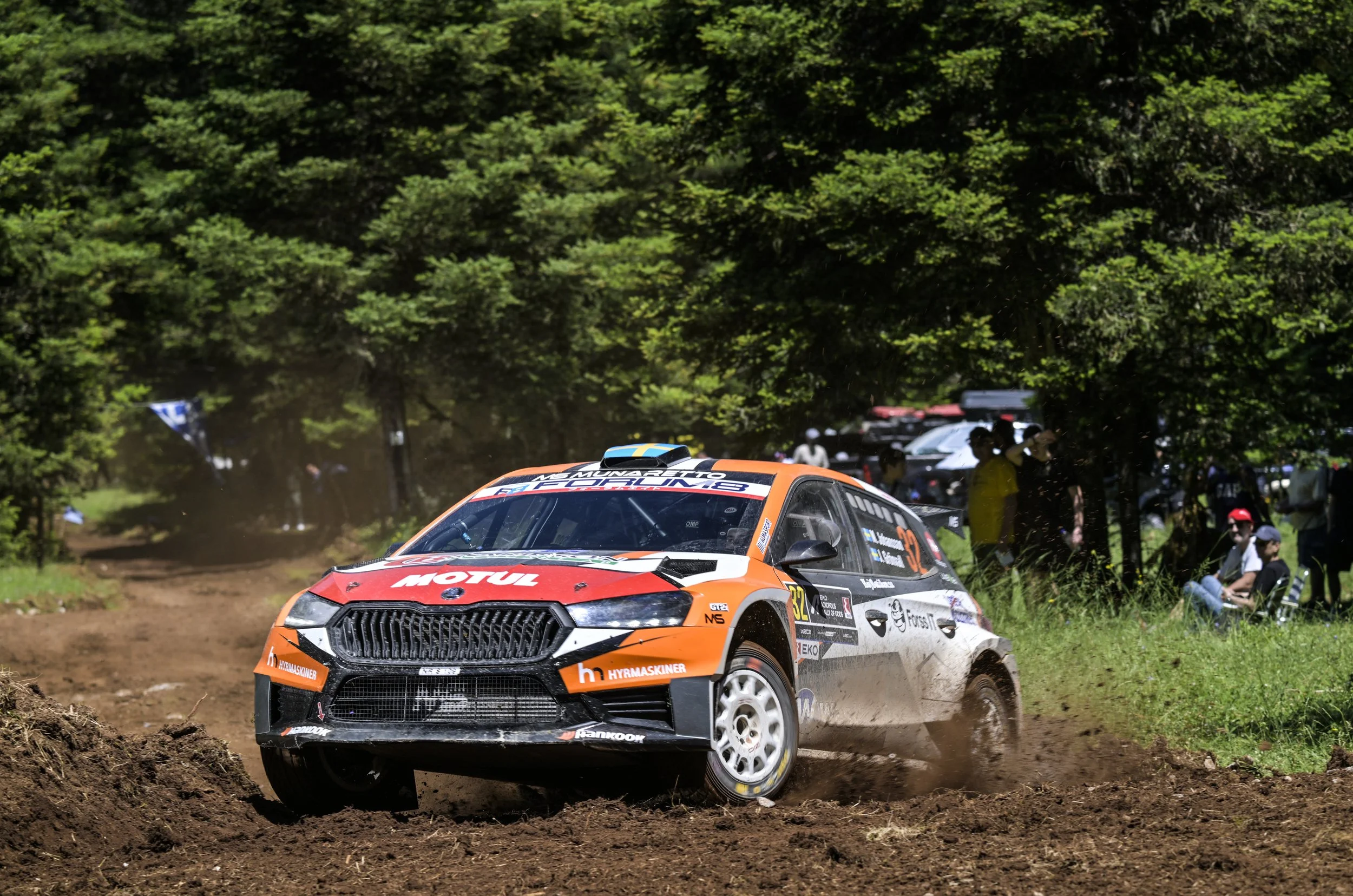 Rally car kicking up dirt on a forested rally stage, with spectators watching from the side.