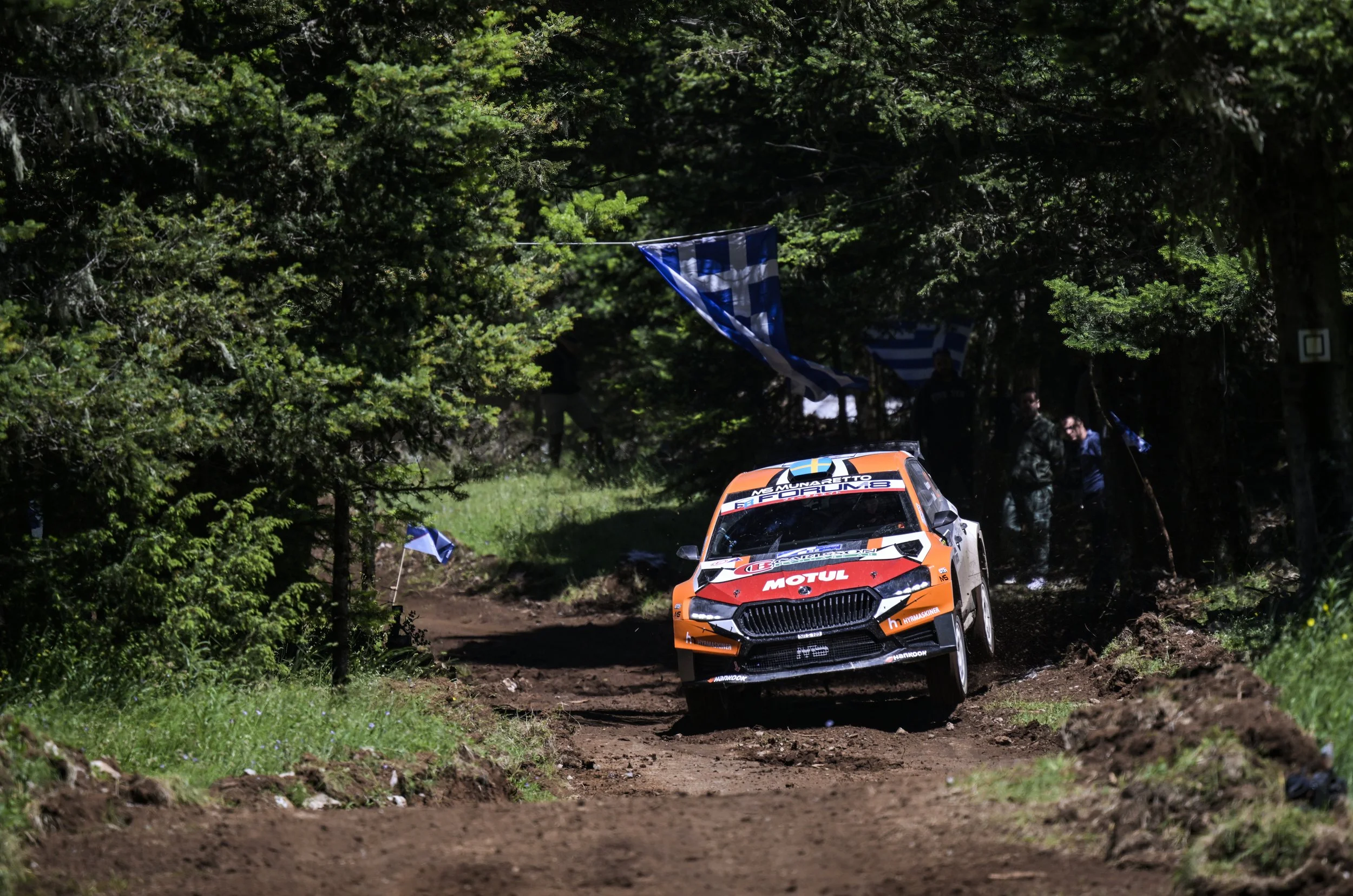 A rally car racing on a dirt trail through a forest with green trees and flags along the course.