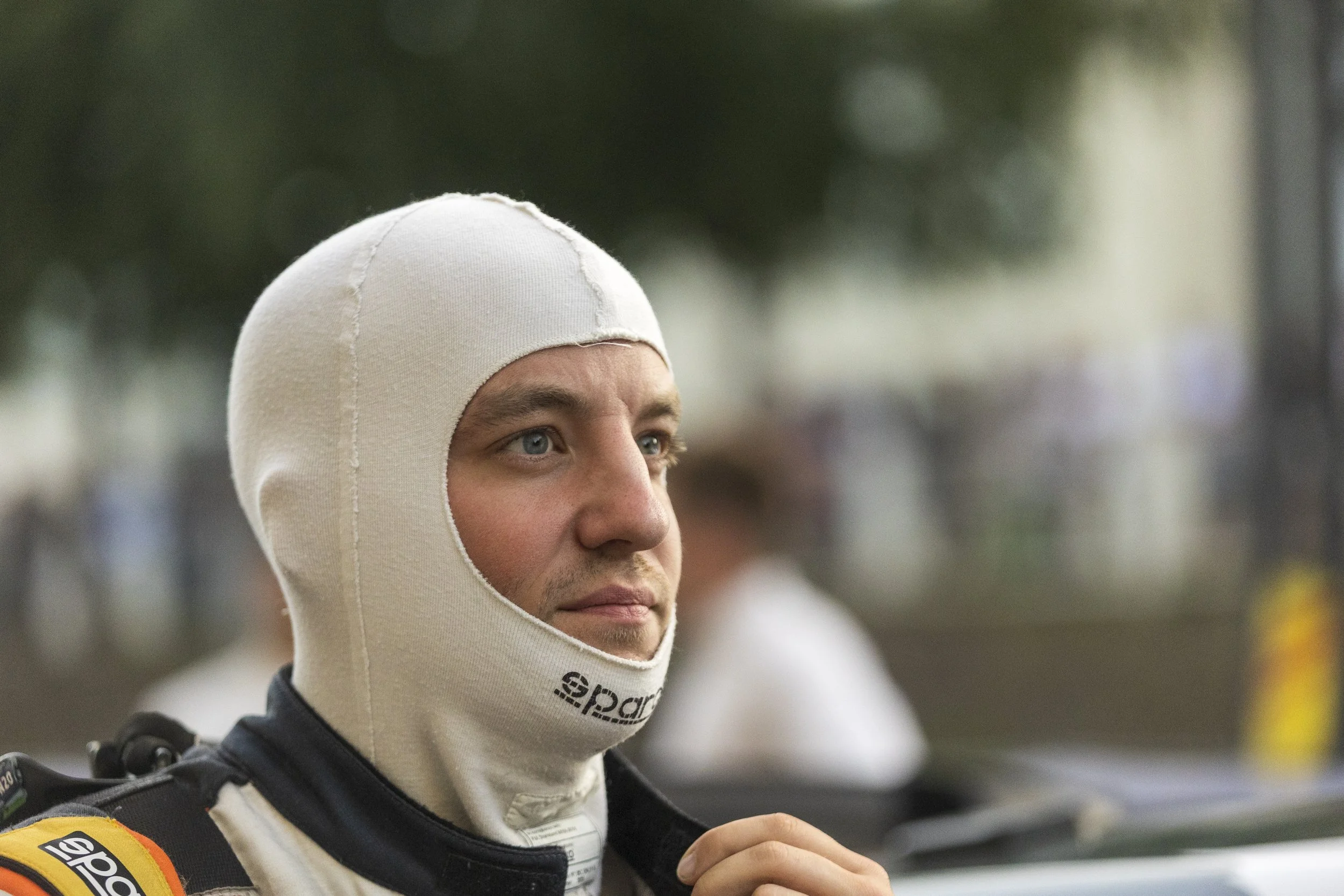 Close-up of a race car driver wearing a white balaclava and a racing suit with yellow, orange, and black patches, standing outdoors.
