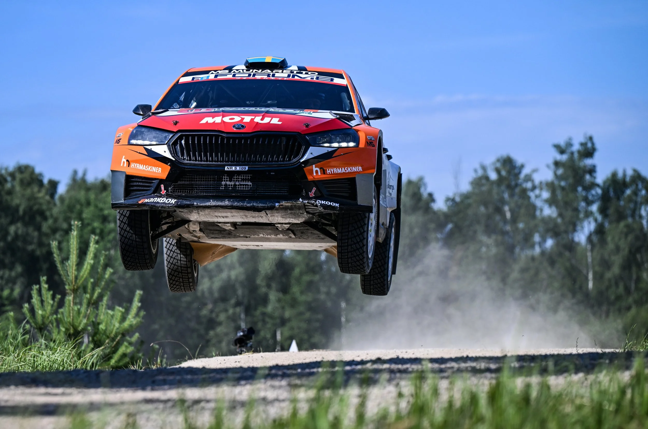 A rally car flying through the air during a race on a dirt road with a clear blue sky and trees in the background.